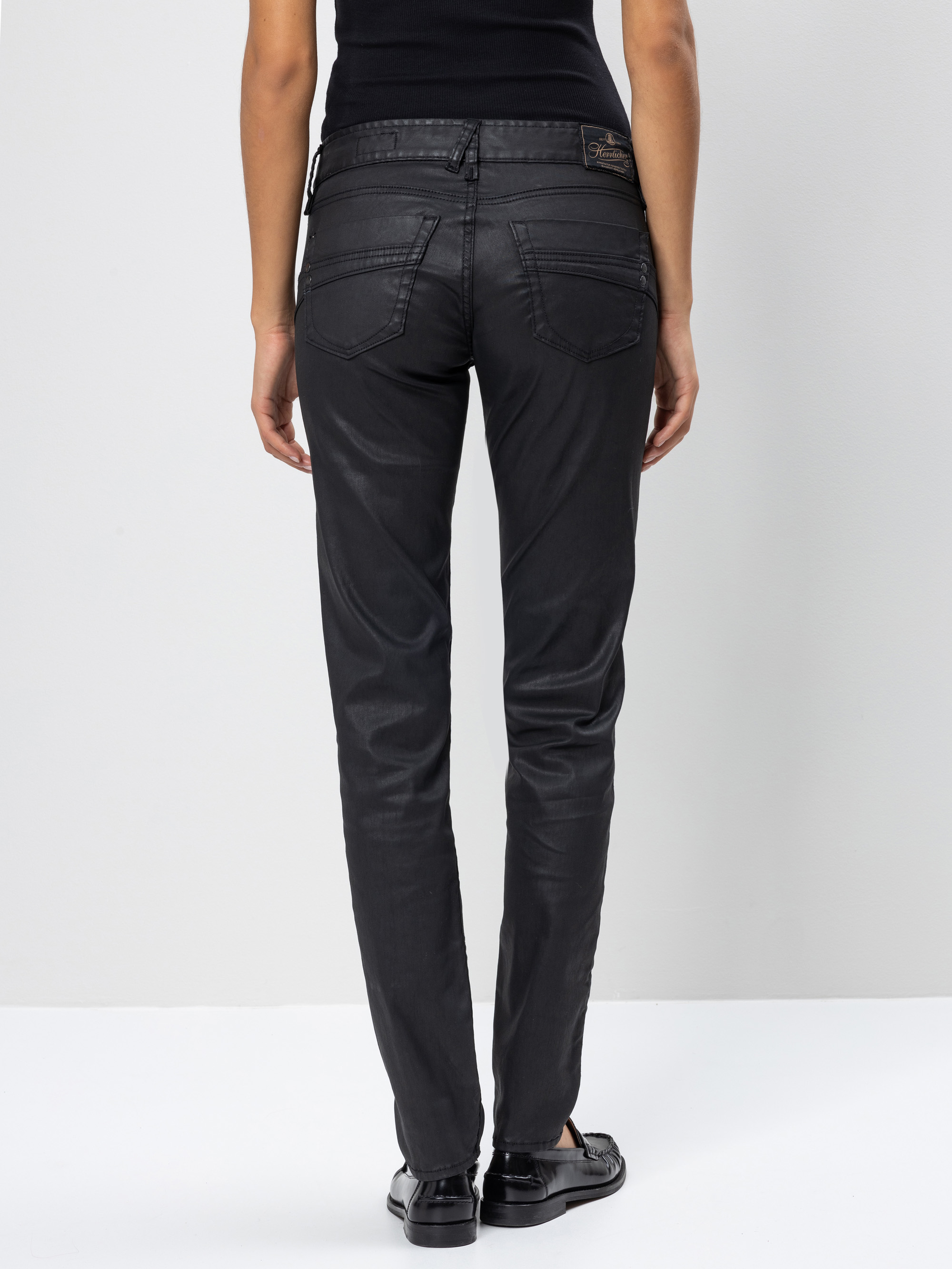 Person in schwarzer Hose mit geradem Bein, schwarzem Oberteil und schwarzen Halbschuhen, die mit dem Rücken zur Kamera vor einem schlichten weißen Hintergrund steht.