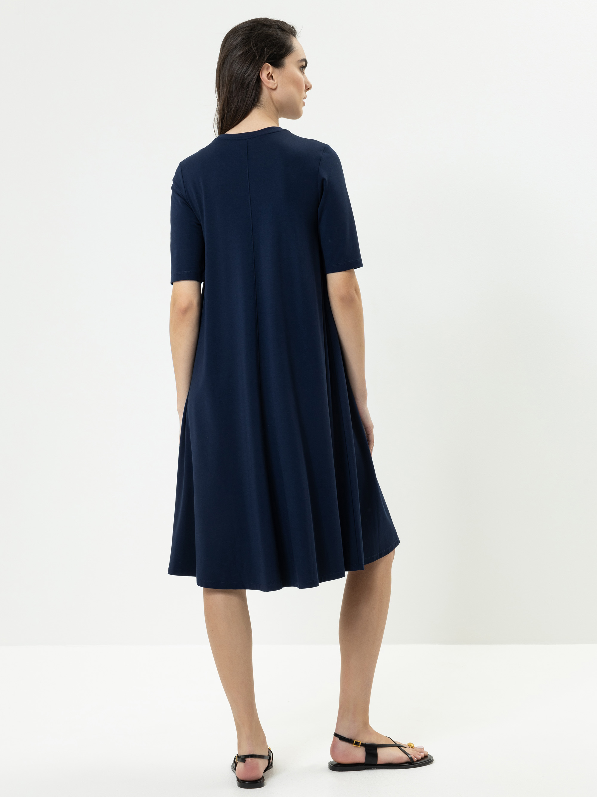 Eine Frau steht mit dem Gesicht zur Wand und trägt ein knielanges, marineblaues, kurzärmeliges Kleid und schwarze Sandalen. Ihr dunkles Haar fällt ihr in den Rücken. Der Hintergrund ist einfarbig weiß.