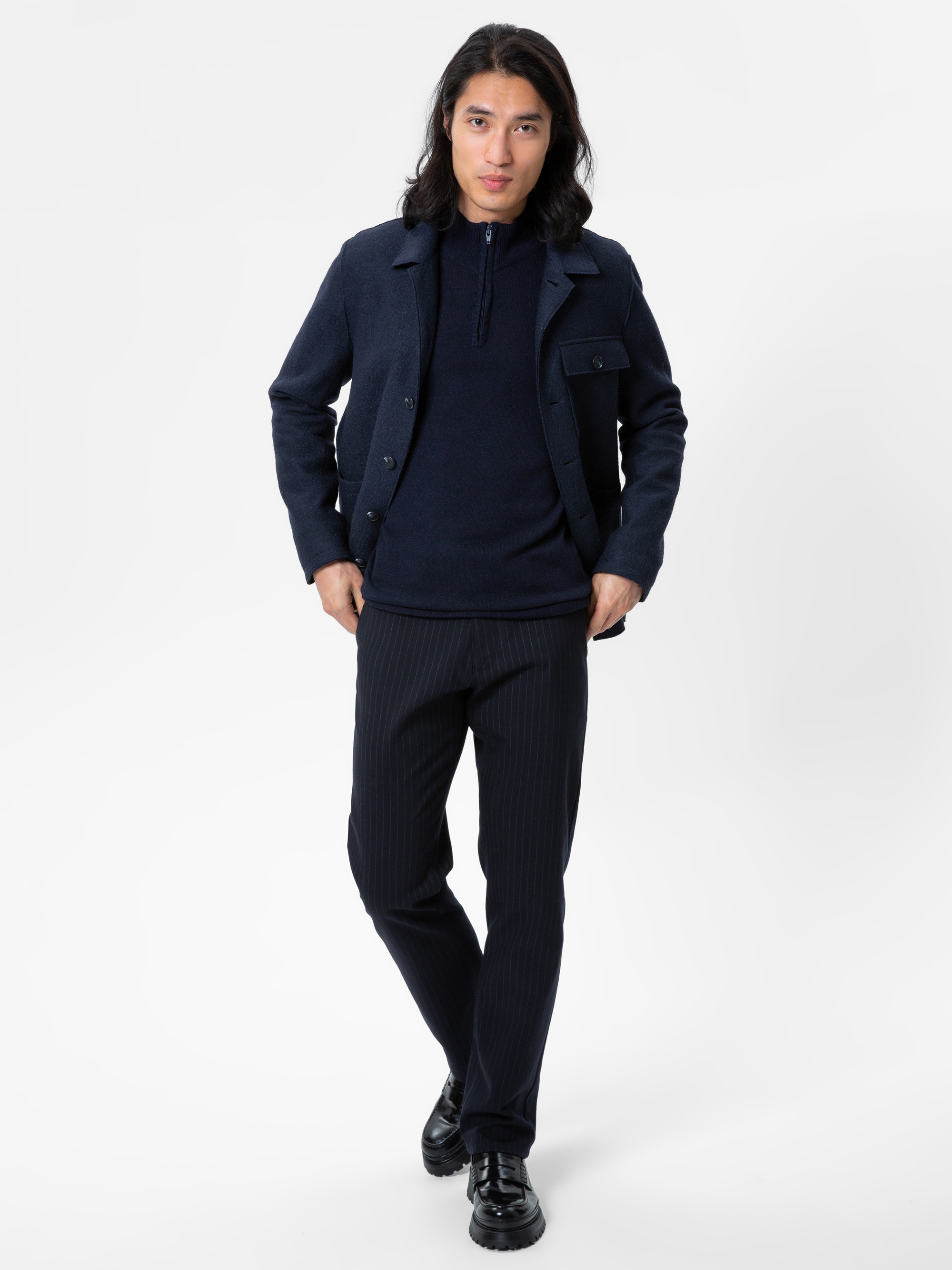 Ein Mann mit langen dunklen Haaren trägt eine marineblaue Jacke über einem dunklen Pullover und eine Hose mit Nadelstreifen. Er steht mit den Händen in den Taschen vor einem schlichten weißen Hintergrund und blickt nach vorne.