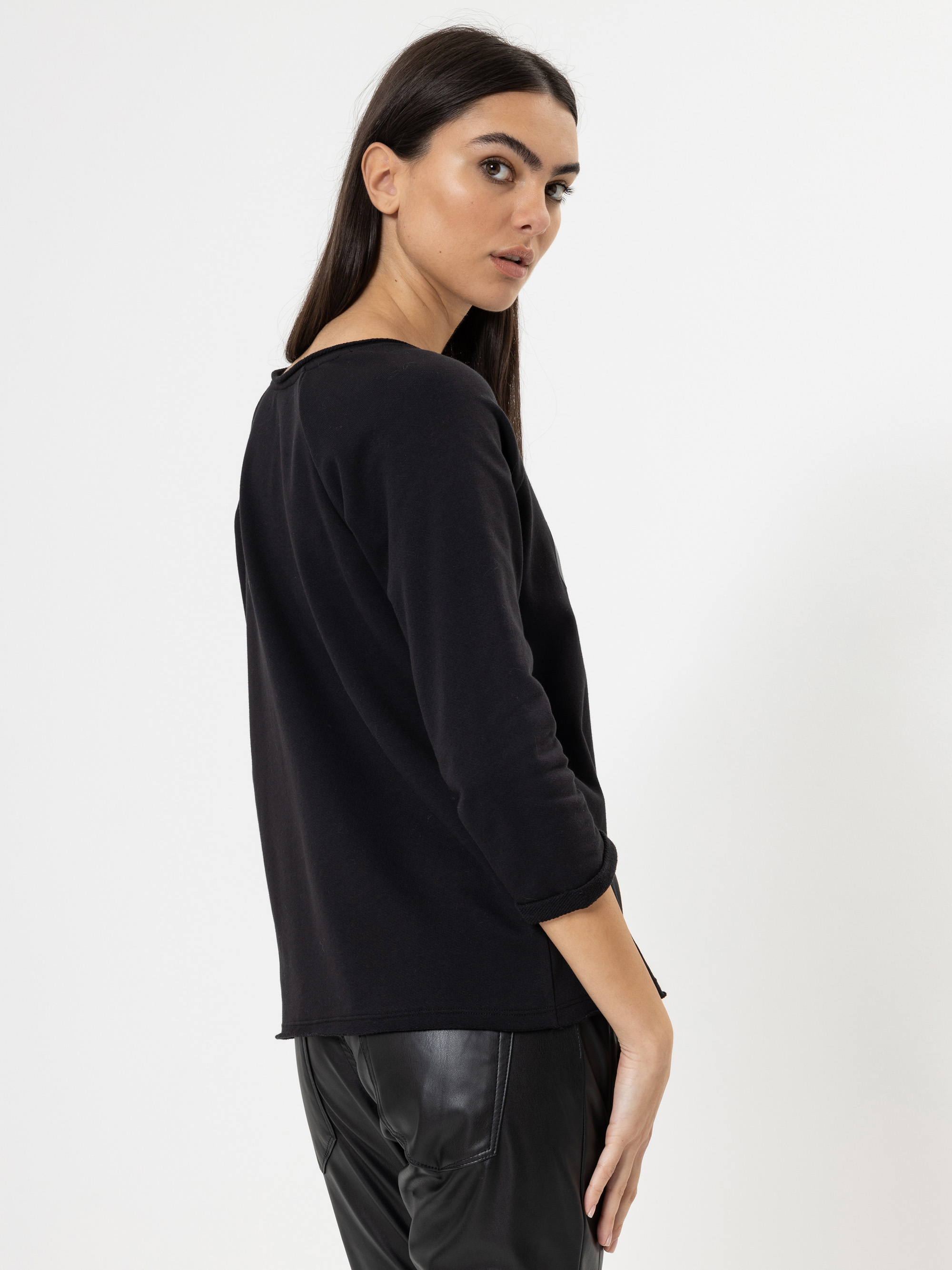 Eine Frau mit langen dunklen Haaren, die ein schwarzes langärmeliges Oberteil und eine schwarze Lederhose trägt, steht vor einem schlichten weißen Hintergrund und schaut über ihre Schulter in die Kamera.