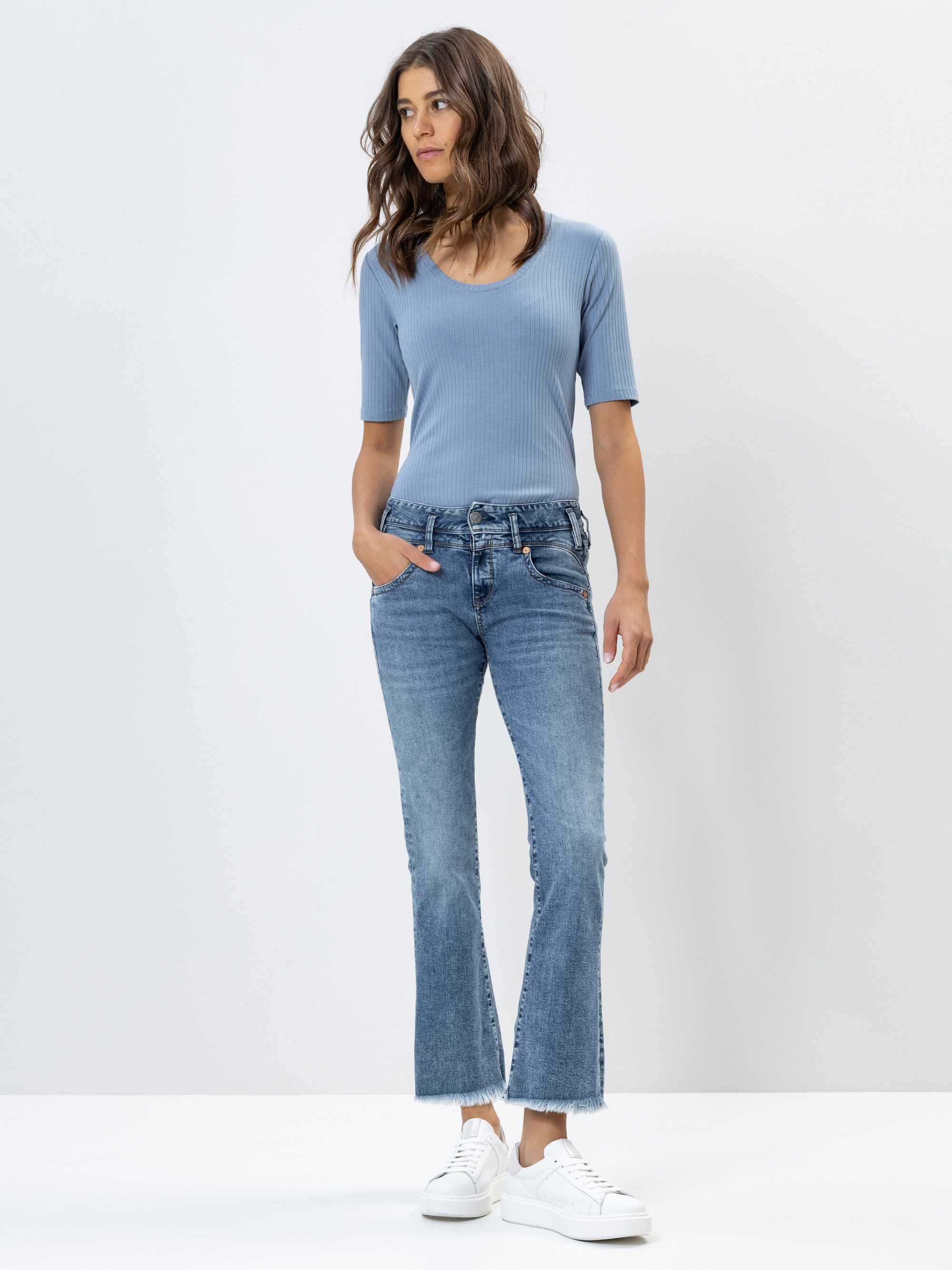 Frau, die vor einem schlichten, hellen Hintergrund steht, ein hellblaues, kurzärmeliges, geripptes Oberteil, blaue Schlagjeans und weiße Turnschuhe trägt, eine Hand in der Tasche hat und zur Seite schaut.