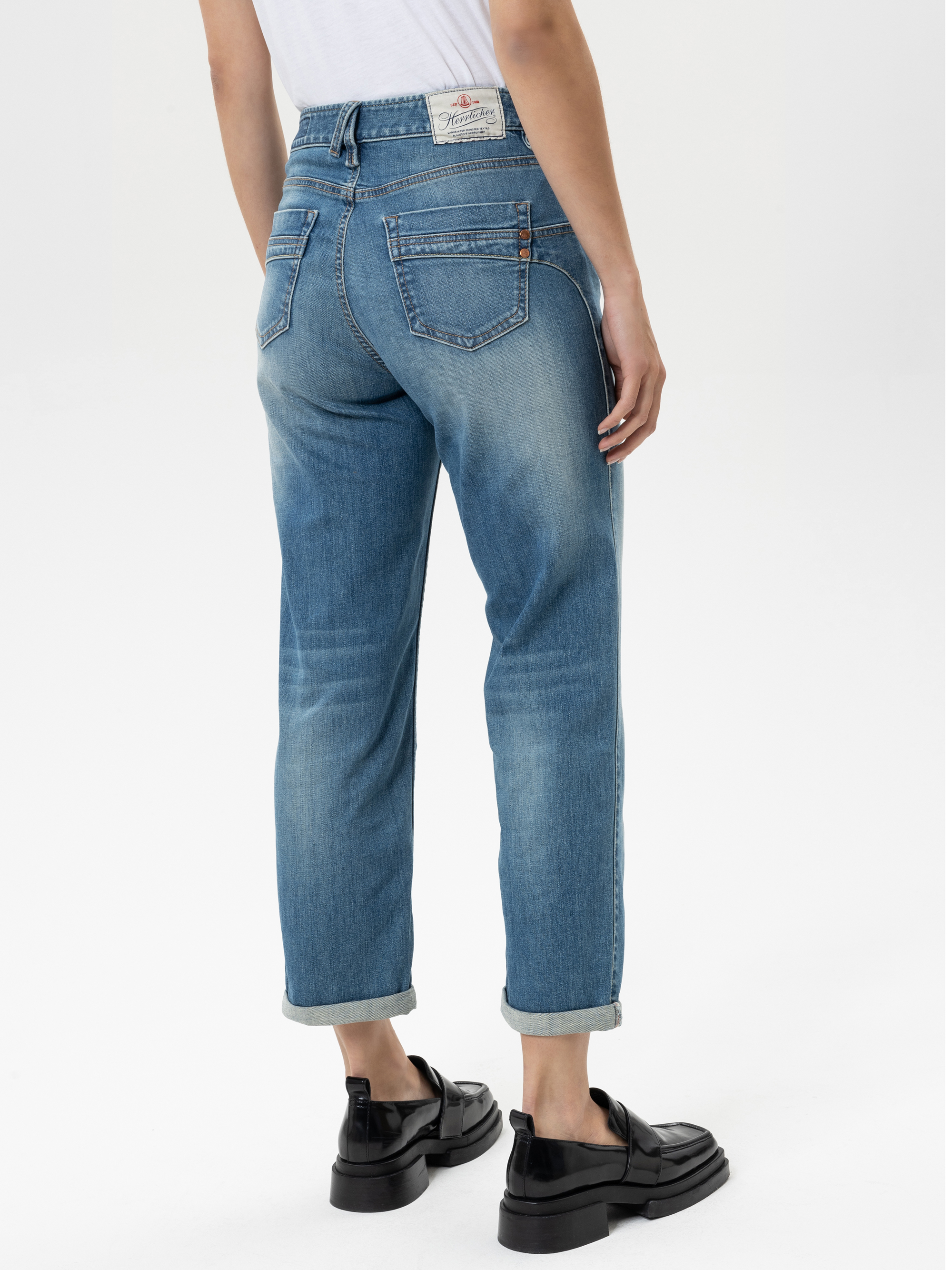 Person, die hellblaue Jeans mit Manschetten, ein weißes T-Shirt und schwarze klobige Halbschuhe trägt und mit dem Rücken zur Kamera vor einem schlichten hellen Hintergrund steht.