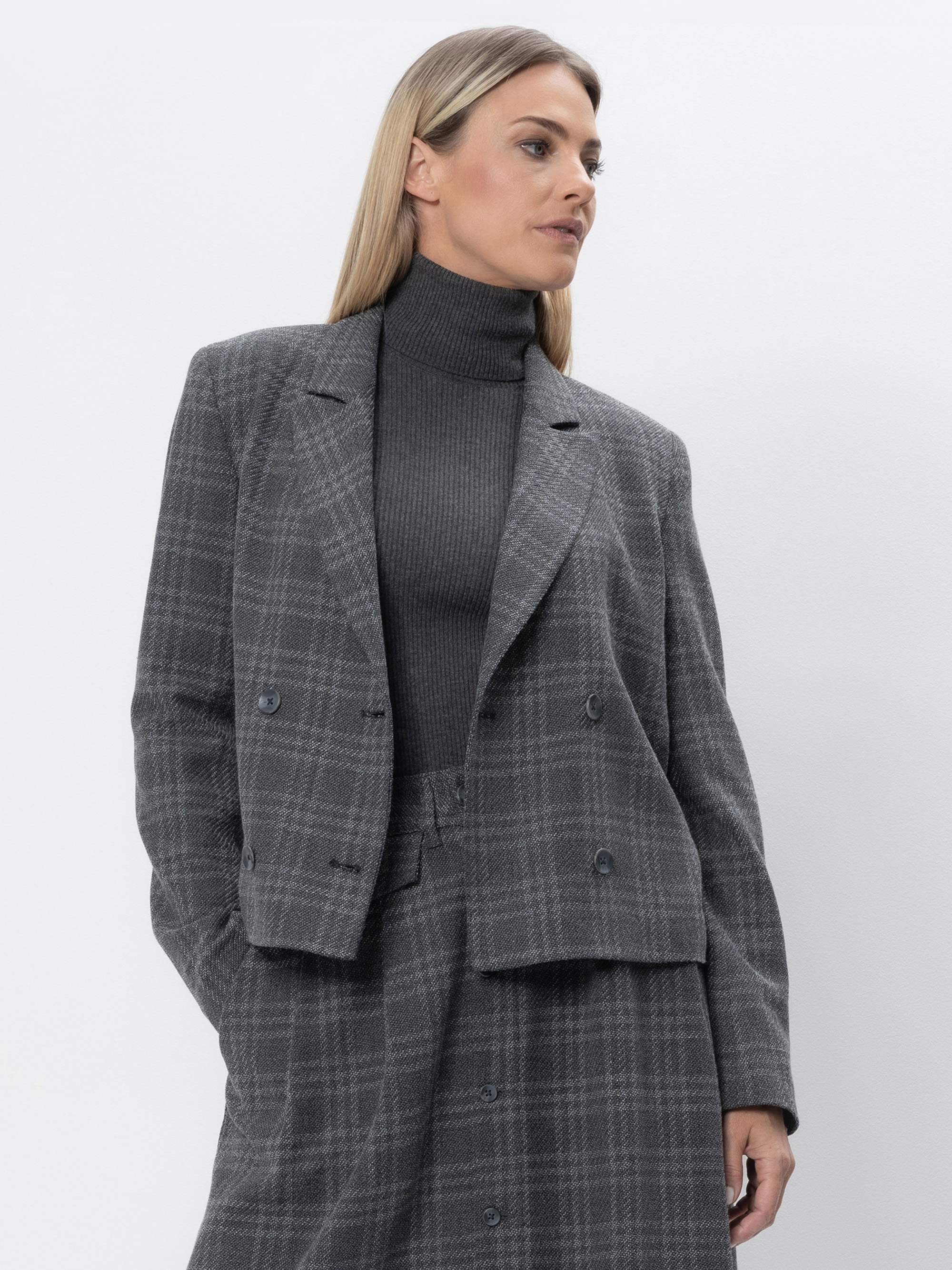 Eine Frau mit glattem, blondem Haar trägt eine grau karierte Anzugsjacke und einen dazu passenden Rock über einem dunkelgrauen, gerippten Rollkragenpullover. Sie steht vor einem schlichten weißen Hintergrund und blickt nach links.