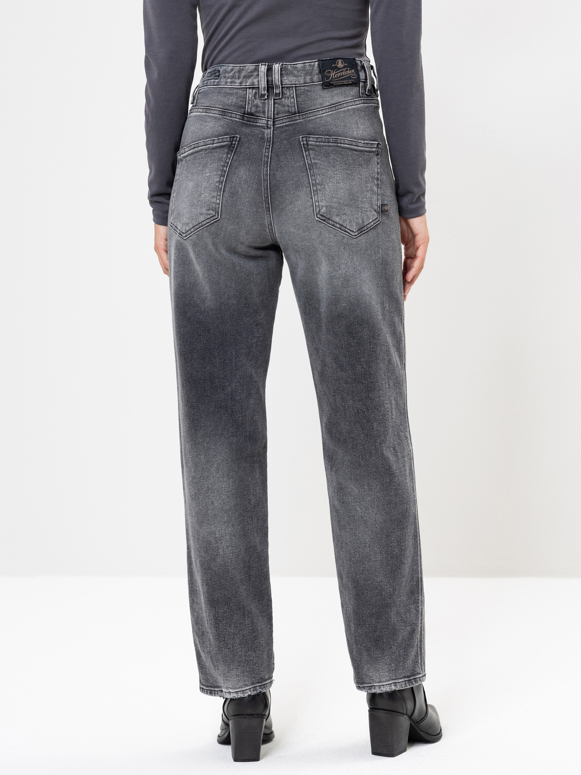 Eine Person, die hochgeschnittene schwarze Jeans mit geradem Bein und schwarzen Stiefeln mit Absatz trägt, wird von hinten gezeigt und steht vor einem schlichten weißen Hintergrund.