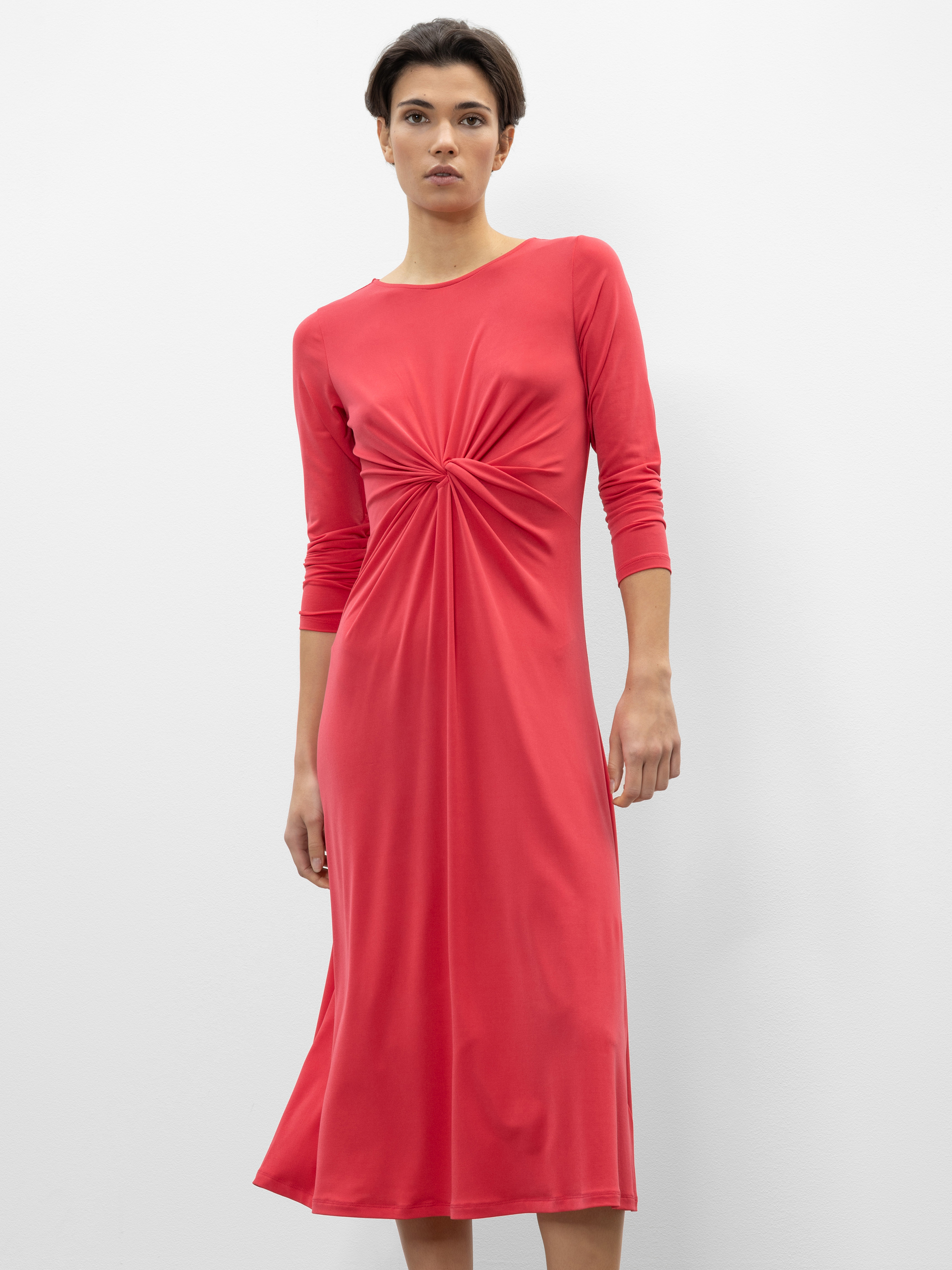 Eine Person, die ein langärmeliges, knielanges rotes Kleid mit einem gerafften Knoten in der Taille trägt, steht vor einem schlichten weißen Hintergrund.