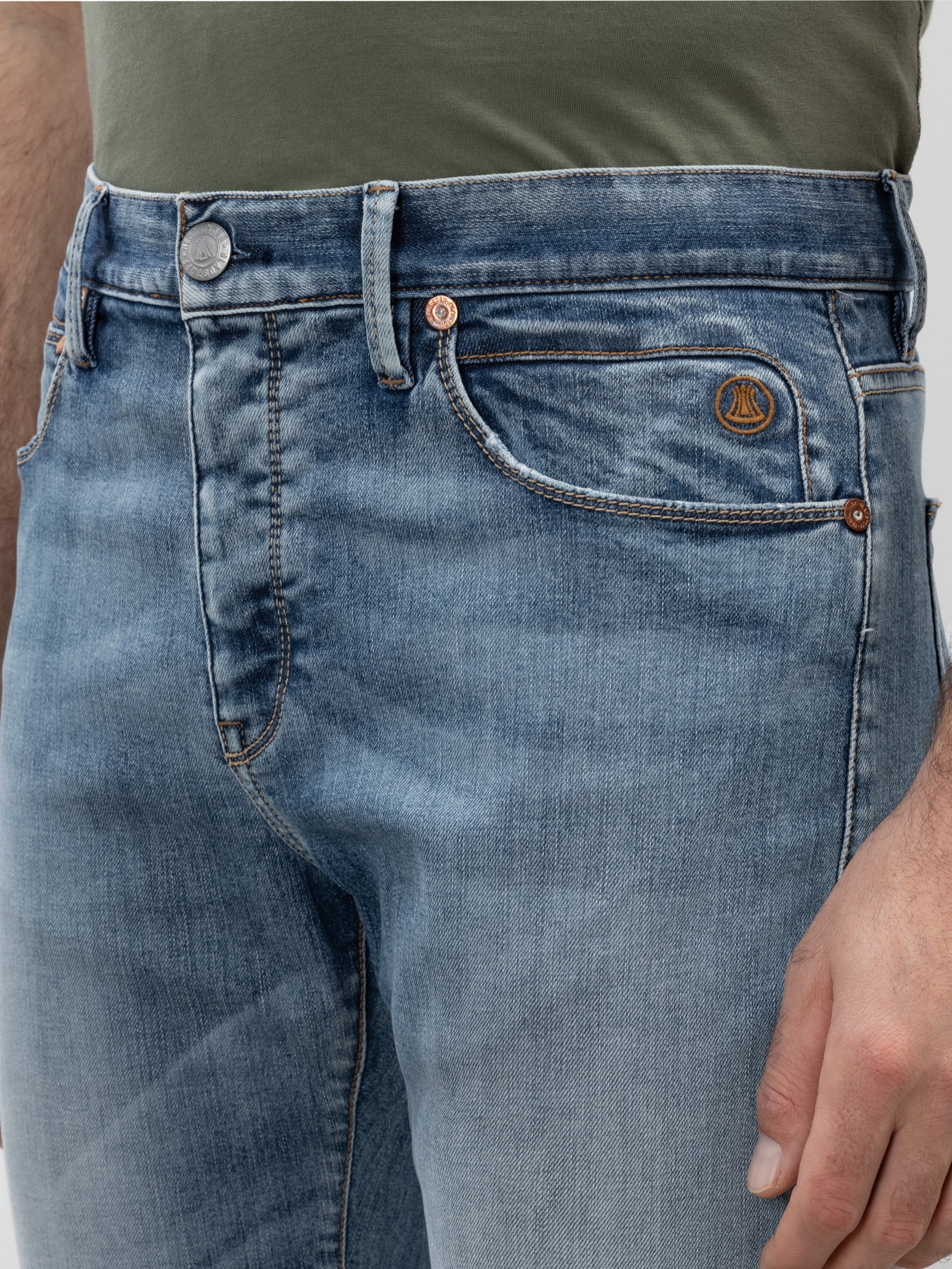 Eine Person, die eine hellblaue Jeans und ein olivgrünes Hemd trägt, das von der Taille bis zur Mitte des Oberschenkels reicht und ein sichtbares Logo auf der kleinen Vordertasche aufweist.