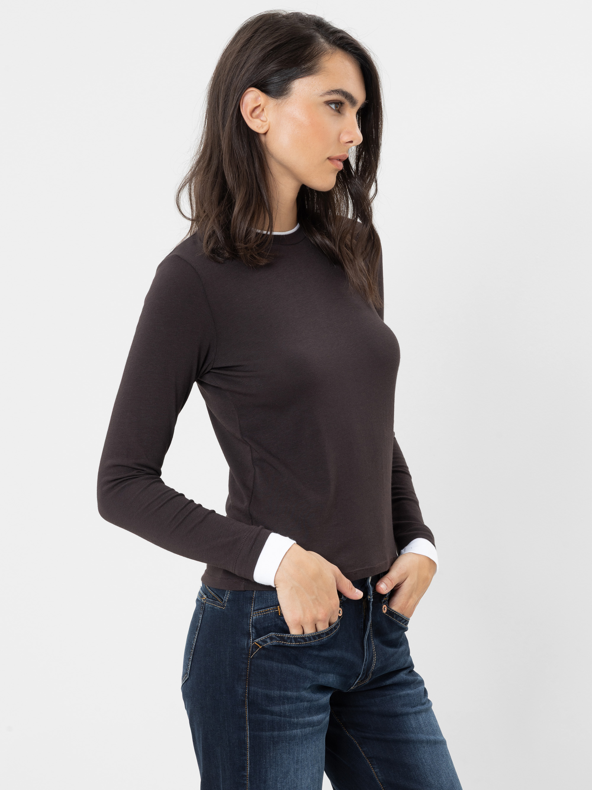 Frau mit langen dunklen Haaren, die ein dunkles Langarmshirt und blaue Jeans trägt, in einer entspannten Haltung steht, eine Hand in der Hosentasche hat und vor einem schlichten weißen Hintergrund nach rechts blickt.