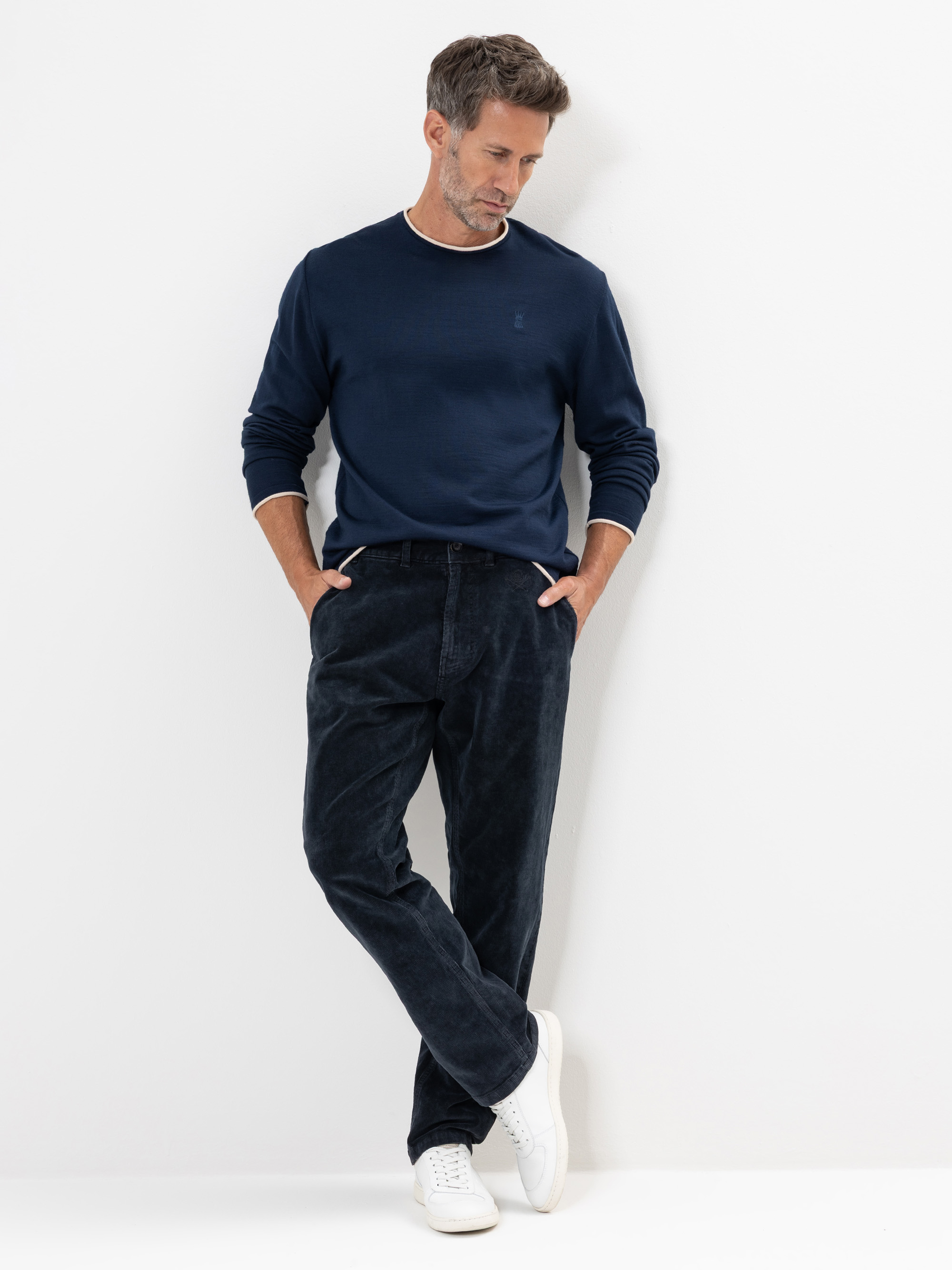 Ein Mann mit kurzen braunen Haaren, einem marineblauen langärmeligen Hemd, einer dunklen Cordhose und weißen Turnschuhen steht an einer weißen Wand und schaut mit den Händen in den Taschen nach unten.