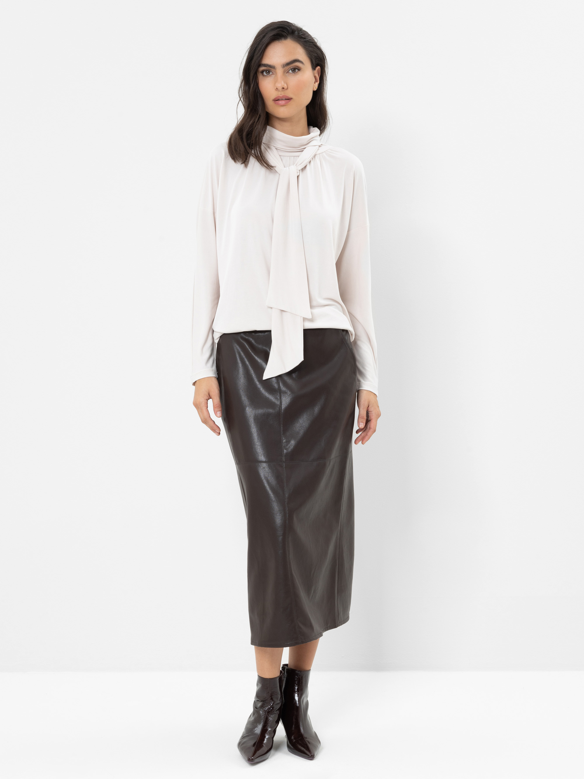 Eine Frau steht vor einem weißen Hintergrund und trägt eine helle, langärmelige Bluse mit Schleife, einen schwarzen Leder-Midirock und schwarze Stiefeletten. Sie hat dunkles Haar und einen neutralen Gesichtsausdruck.