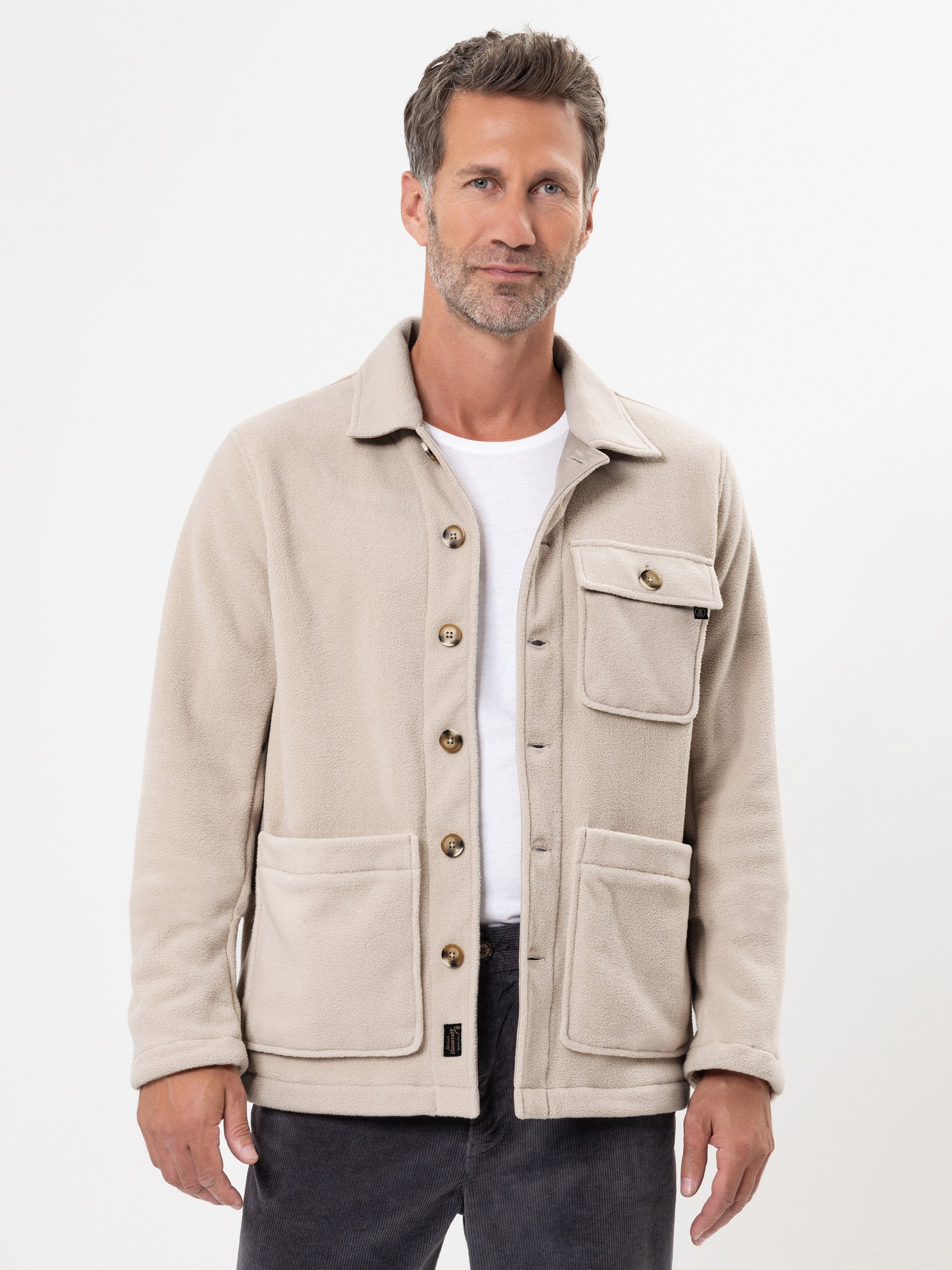 Ein Mann mit kurzen braunen Haaren und gestutztem Bart trägt eine beigefarbene Fleecejacke mit Knopfleiste über einem weißen T-Shirt und einer dunklen Hose. Er steht vor einem schlichten weißen Hintergrund und schaut mit einem leichten Lächeln in die