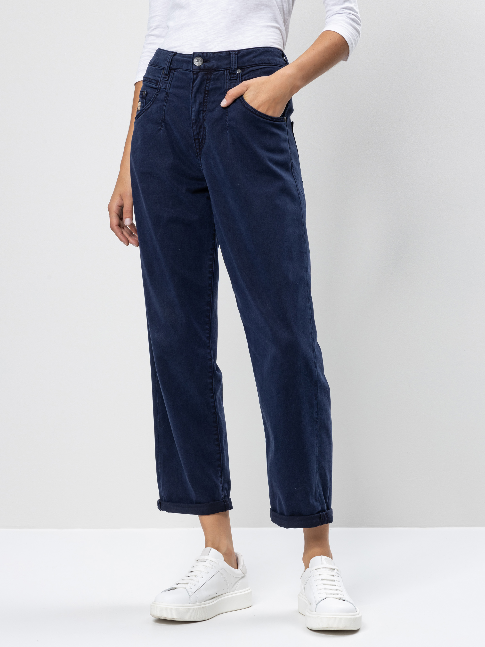 Person, die eine marineblaue Hose mit Manschetten, ein weißes langärmeliges Oberteil und weiße Turnschuhe trägt und mit einer Hand in der Tasche vor einem hellen Hintergrund steht.