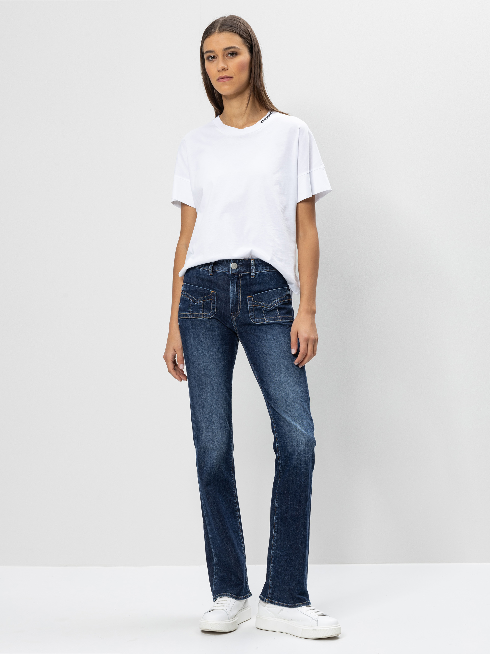 Eine Frau steht vor einem schlichten weißen Hintergrund und trägt ein weißes T-Shirt, blaue Jeans und weiße Turnschuhe. Sie hat glattes braunes Haar und einen neutralen Gesichtsausdruck, ihre Arme liegen entspannt an ihren Seiten.