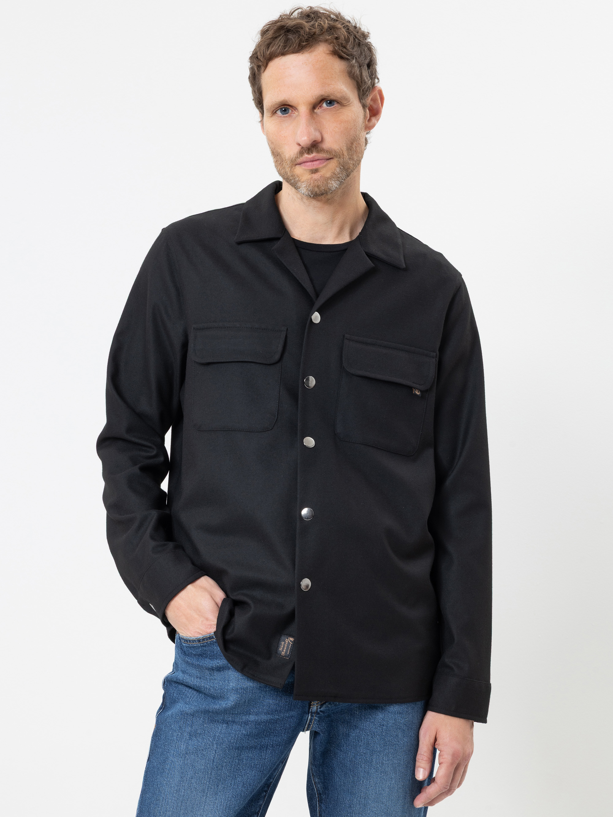 Ein Mann mit kurzen, gewellten Haaren, der ein schwarzes Hemd mit Knöpfen und zwei Brusttaschen sowie eine blaue Jeans trägt, steht vor einem schlichten weißen Hintergrund und hat eine Hand in der Tasche.