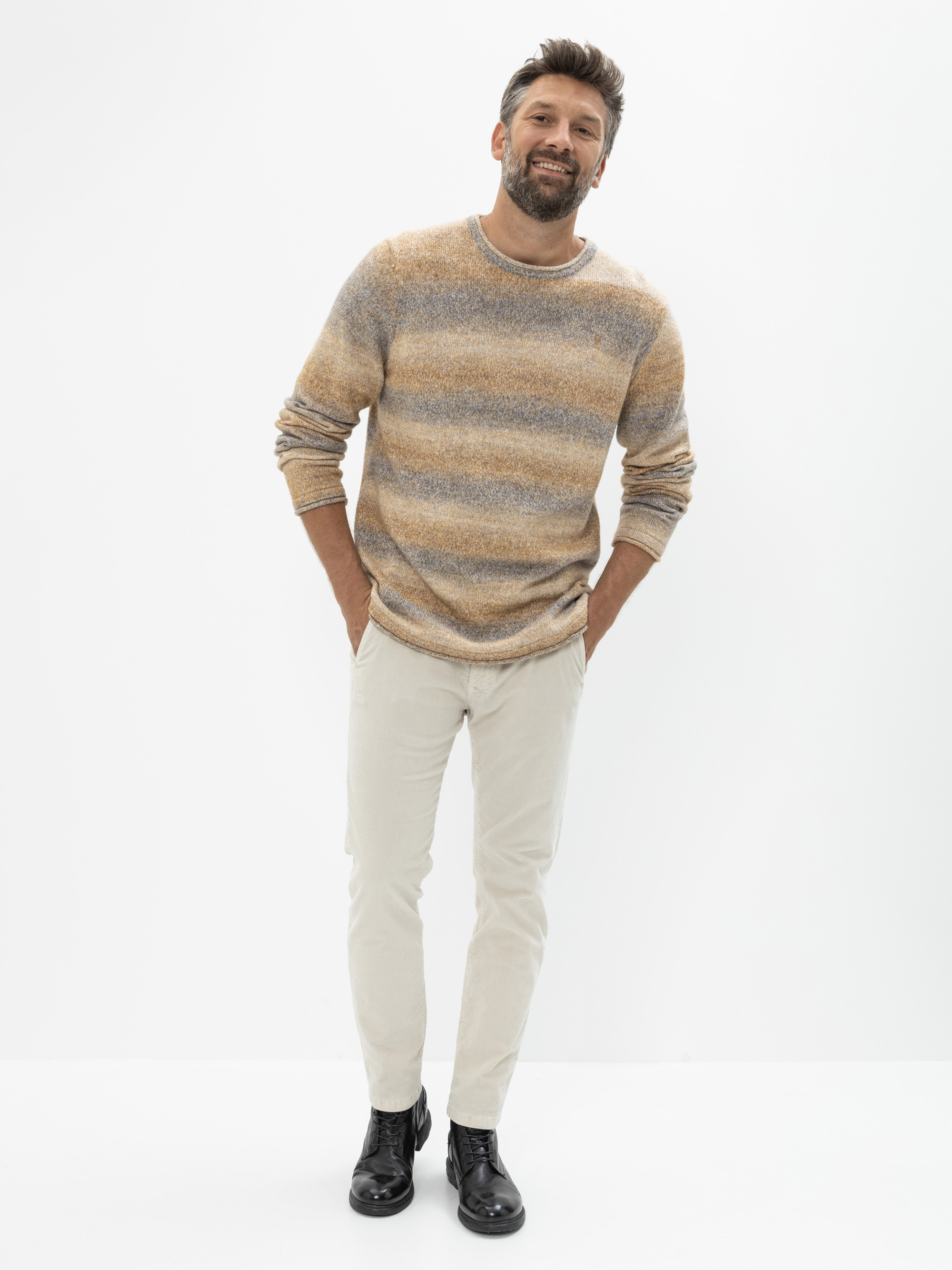 Ein Mann mit kurzen dunklen Haaren und einem Bart steht lächelnd vor einem schlichten weißen Hintergrund. Er trägt einen gestreiften Pullover in Erdtönen, eine helle Hose und schwarze Stiefel, wobei er die Hände in den Taschen hat.