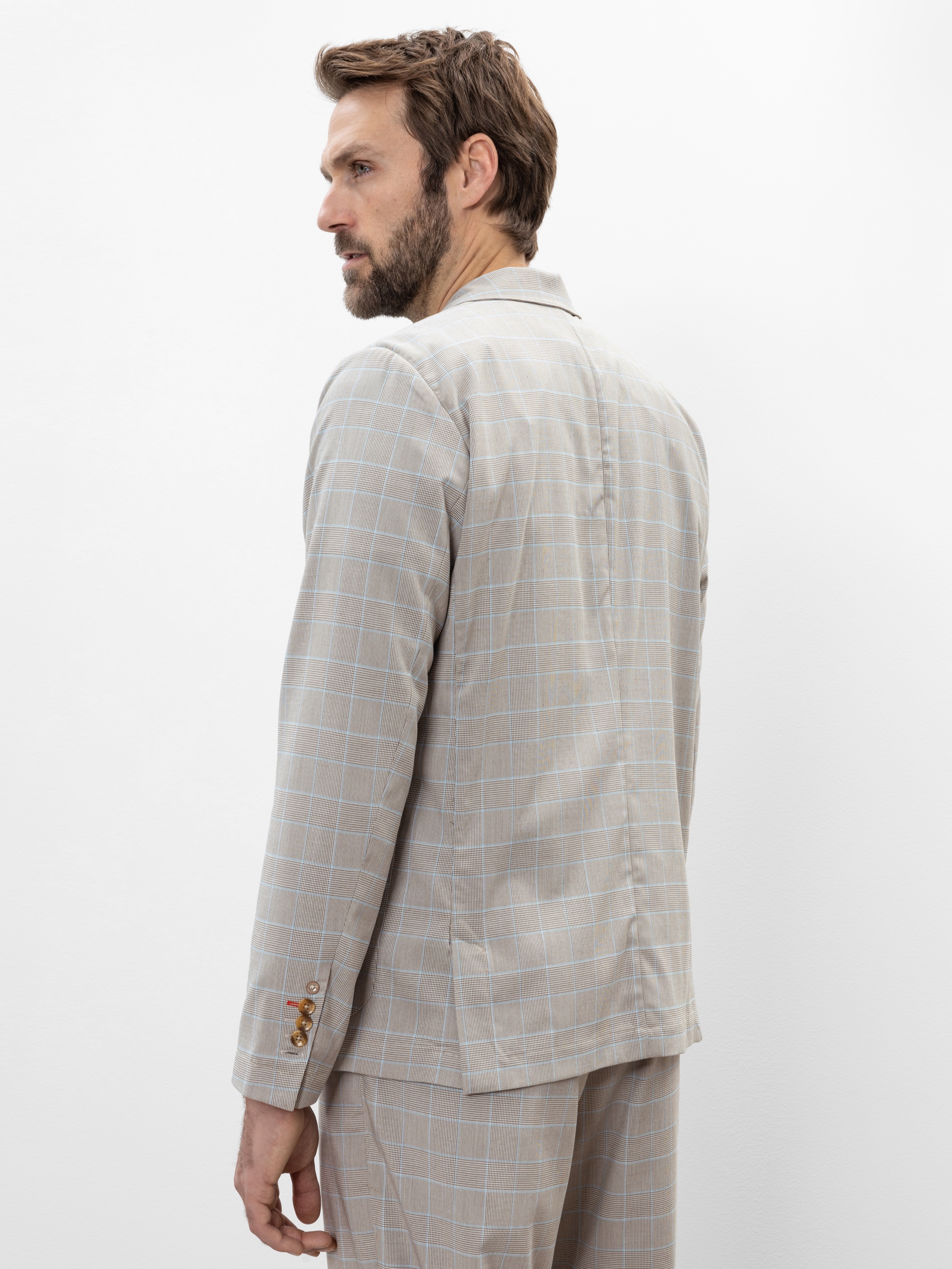 Ein Mann mit hellbraunem Haar und Bart trägt einen hellbeigen karierten Anzug. Er steht seitlich und schaut über seine Schulter vor einem schlichten weißen Hintergrund.