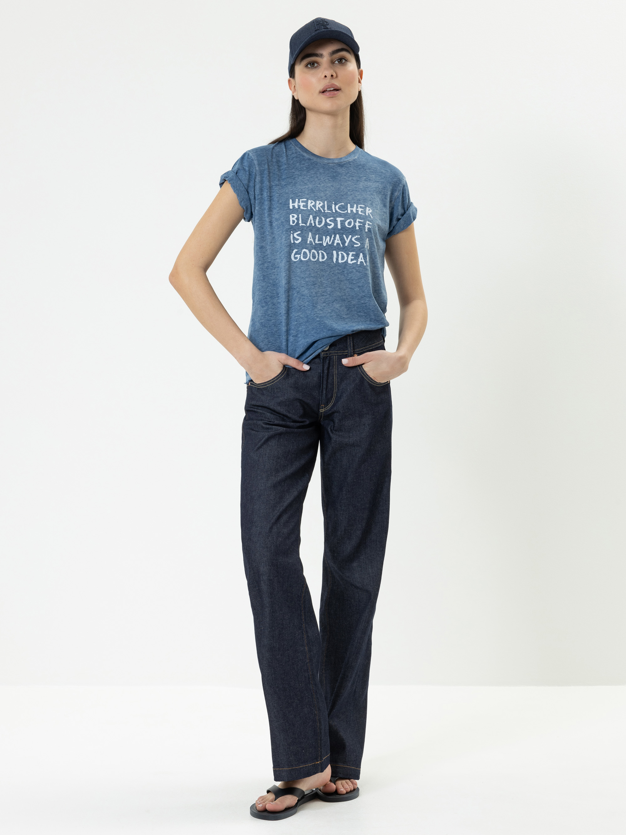 Eine Frau steht vor einem weißen Hintergrund, trägt eine blaue Mütze, ein blaues T-Shirt mit Text, dunkelblaue Jeans und Sandalen, die Hände in den Taschen.