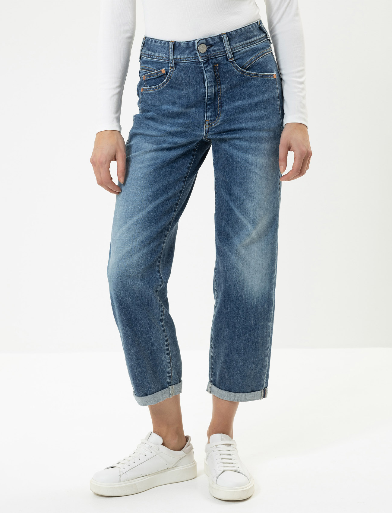 Eine Person, die eine blaue Jeans mit Bündchen, ein langärmeliges weißes Oberteil und weiße Turnschuhe trägt, steht vor einem schlichten weißen Hintergrund. Das Bild konzentriert sich auf die untere Hälfte des Körpers.