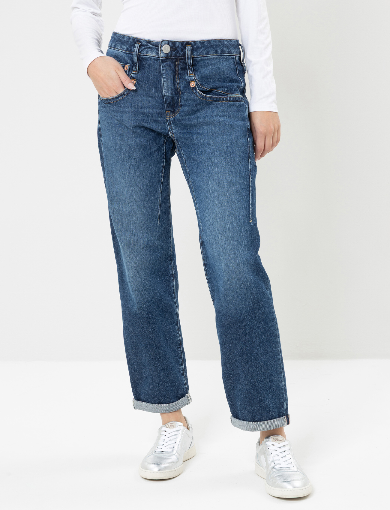 Eine Person, die eine blaue Jeans mit Bündchen, ein langärmeliges weißes Oberteil und weiße Turnschuhe trägt, steht vor einem schlichten weißen Hintergrund. Das Bild konzentriert sich auf die untere Hälfte des Körpers.