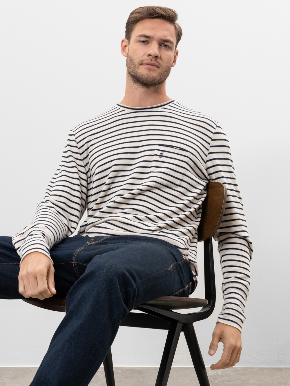 Ein Mann mit kurzen Haaren und gestutztem Bart sitzt auf einem Stuhl, trägt ein weiß-schwarz gestreiftes langärmeliges Hemd und dunkelblaue Jeans vor einem schlichten weißen Hintergrund.