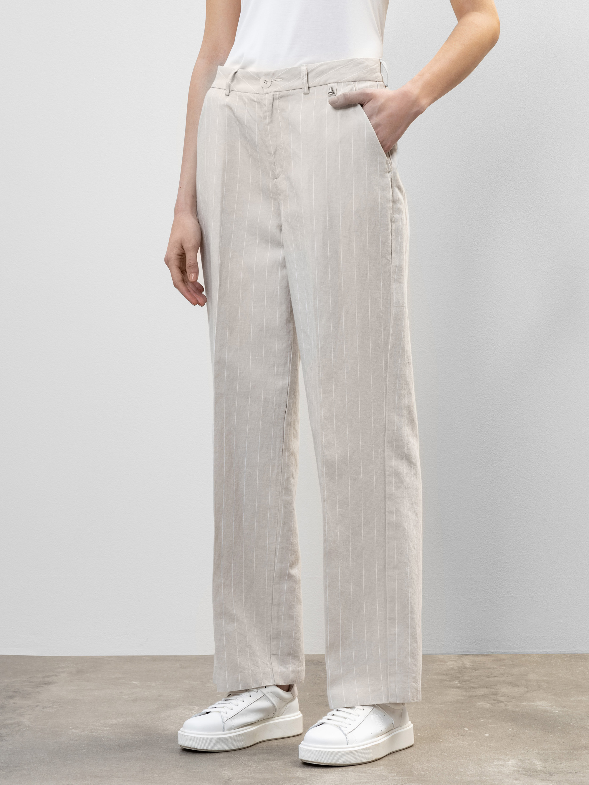 Eine Person, die eine hellbeige, hoch taillierte Hose mit weitem Bein und vertikalen Nadelstreifen, ein weißes T-Shirt und weiße Turnschuhe trägt, steht vor einem schlichten, hellgrauen Hintergrund.