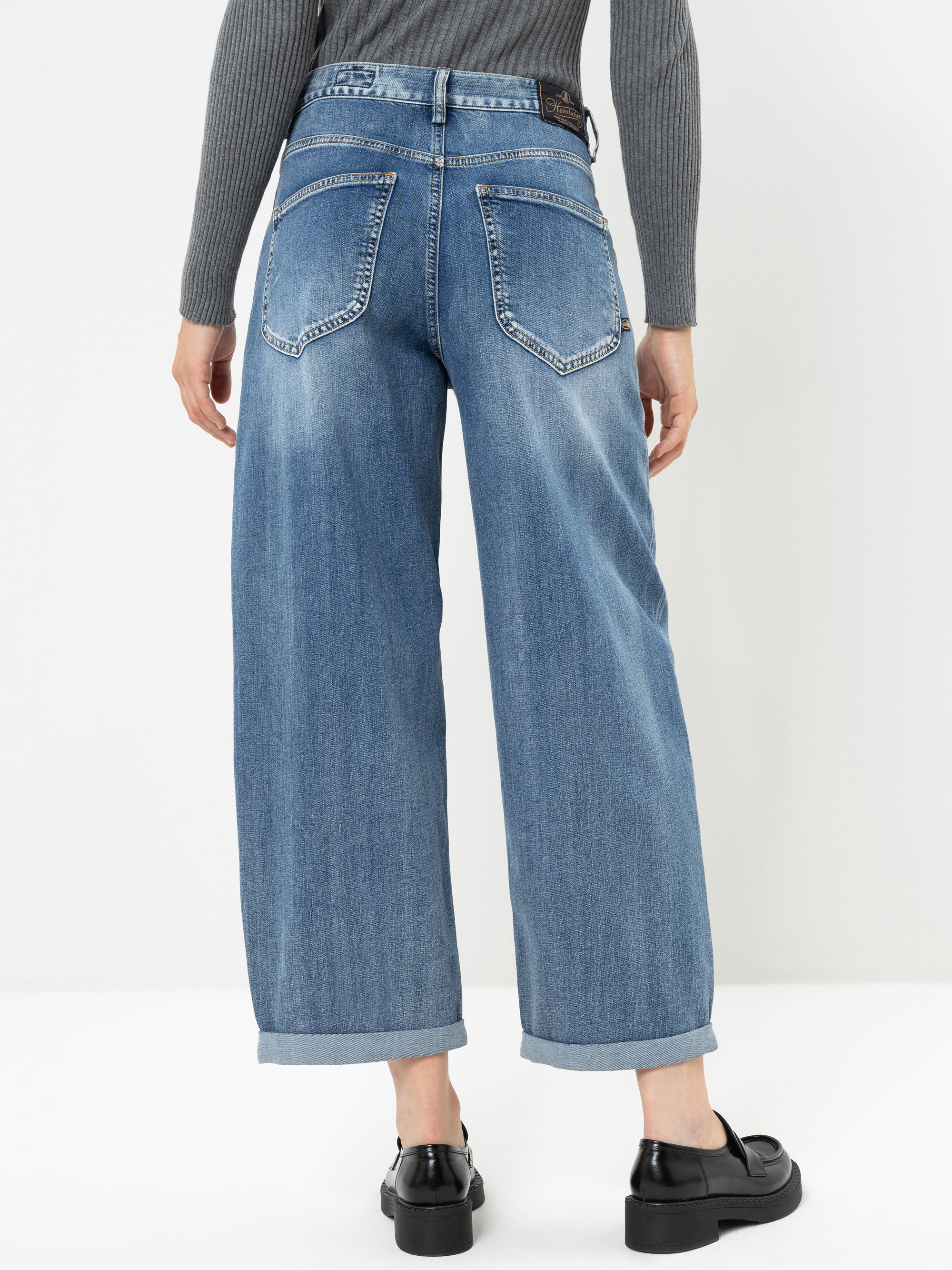 Eine Person, die eine blaue Jeans mit weitem Bein und Umschlag sowie ein graues, geripptes Langarm-Oberteil trägt, von hinten gesehen. Sie steht auf einem weißen Boden und trägt schwarze Slipper. Die Jeans weist sichtbare Nähte und zwei Gesäßtaschen