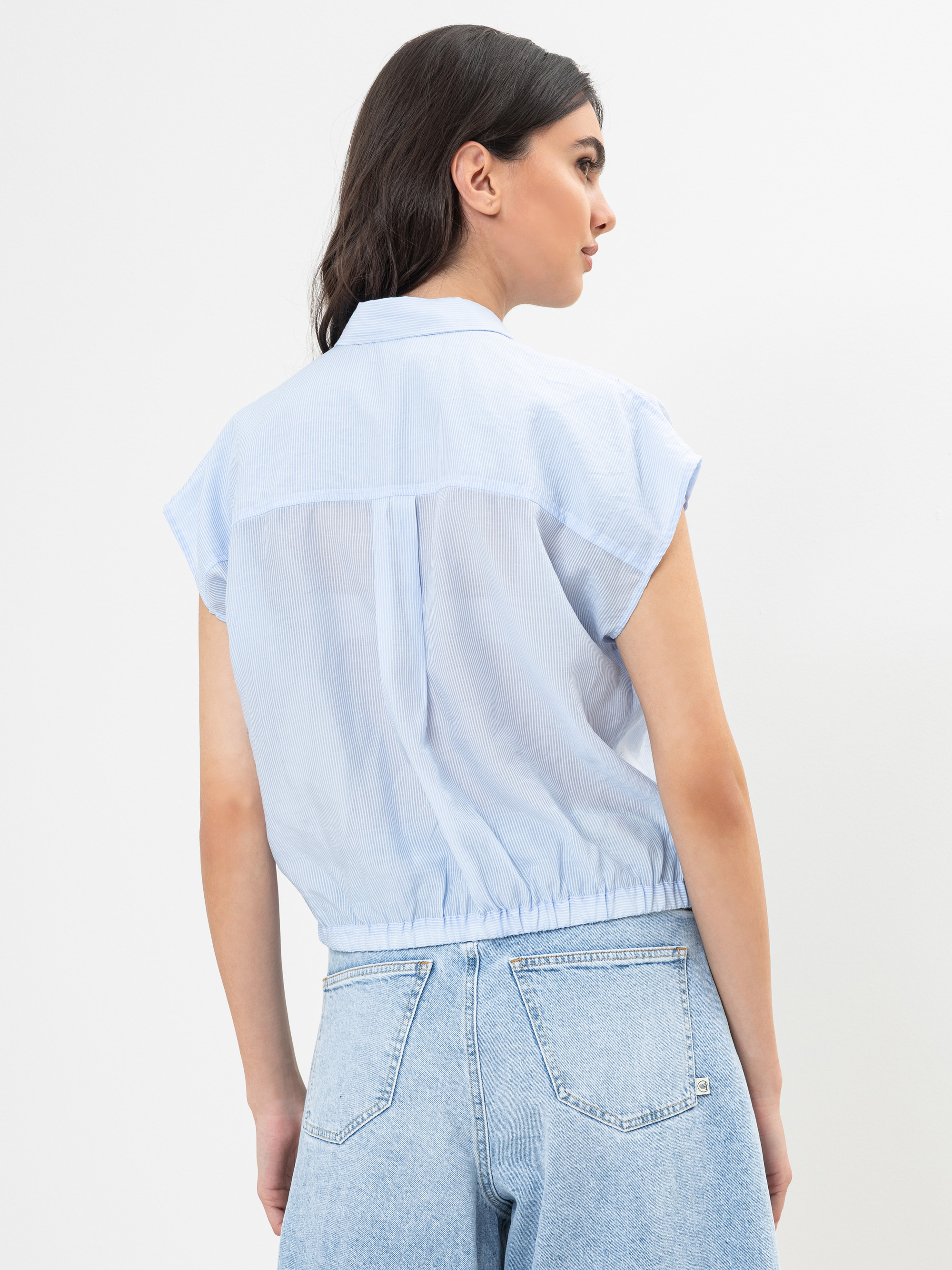 Eine Frau steht mit dem Gesicht zur Wand und trägt eine hellblaue, kurzärmelige Bluse mit Kragen und elastischem Saum, gepaart mit einer hellblauen Jeans mit hoher Taille. Der Hintergrund ist einfarbig weiß.