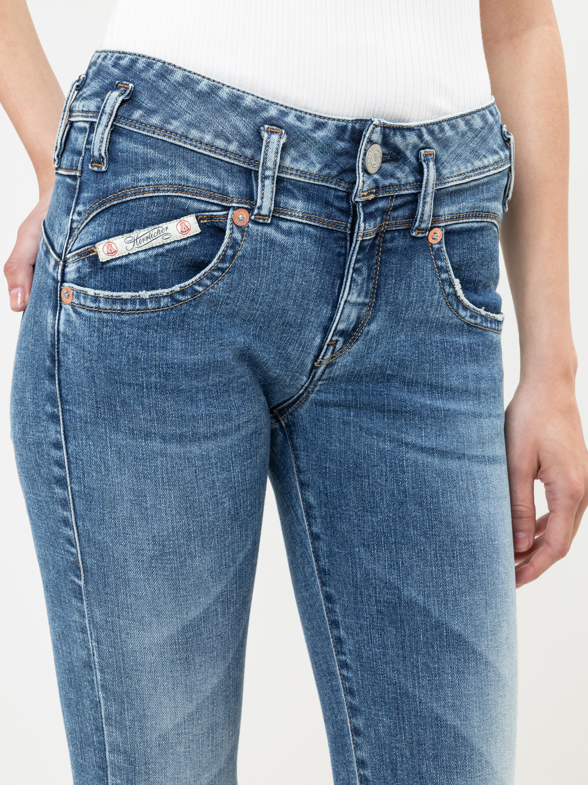 Eine Person, die eine Jeans mittlerer Waschung mit einem sichtbaren Markenetikett auf der Münztasche und ein weißes geripptes Oberteil trägt, von der Taille bis zur Mitte des Oberschenkels, wobei eine Hand in einer Vordertasche ruht.
