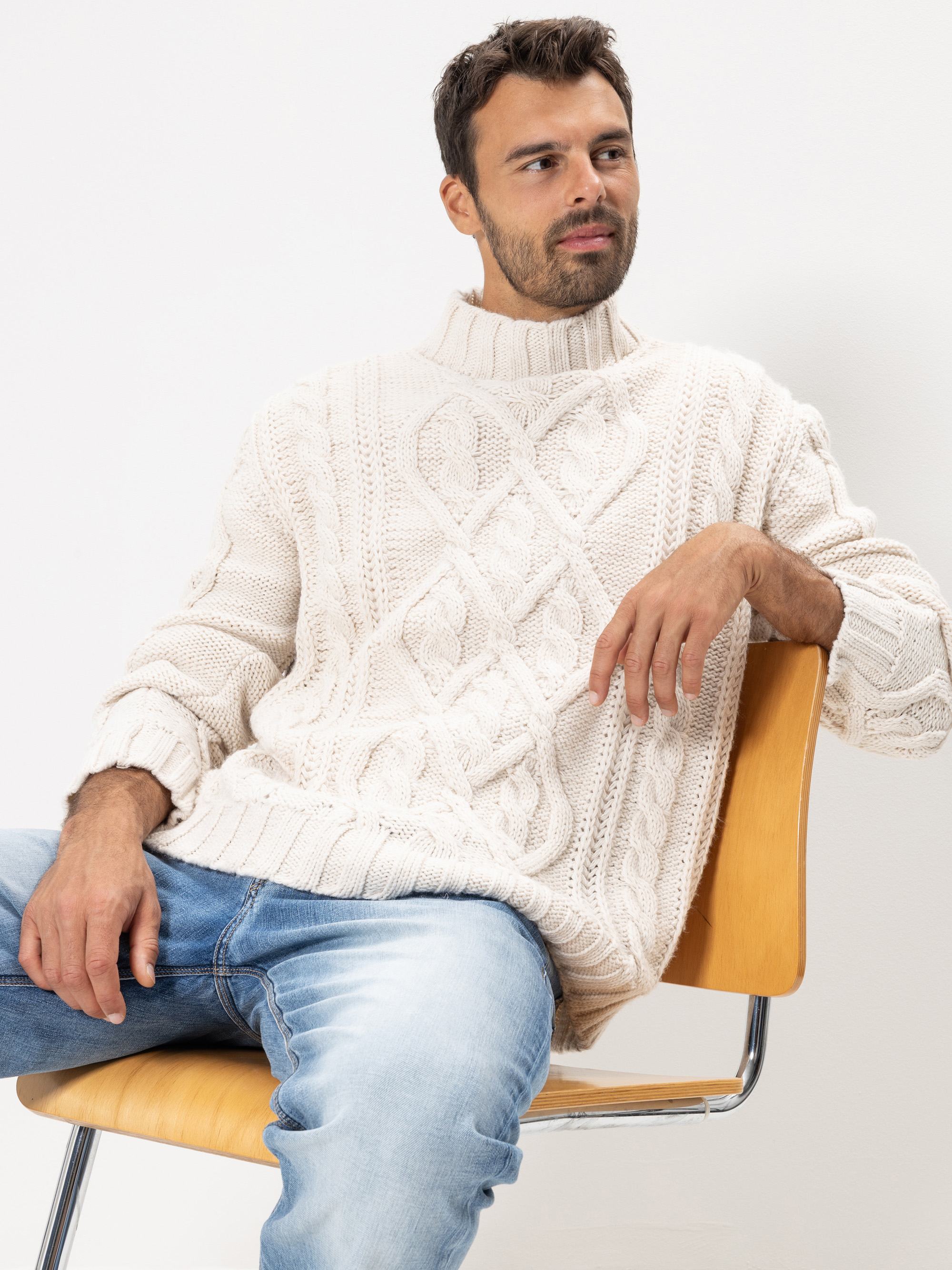 Ein Mann mit kurzen dunklen Haaren und Bart sitzt auf einem Holzstuhl, trägt einen cremefarbenen Strickpullover mit Rautenmuster und hellblaue Jeans und blickt vor einem schlichten hellen Hintergrund zur Seite.