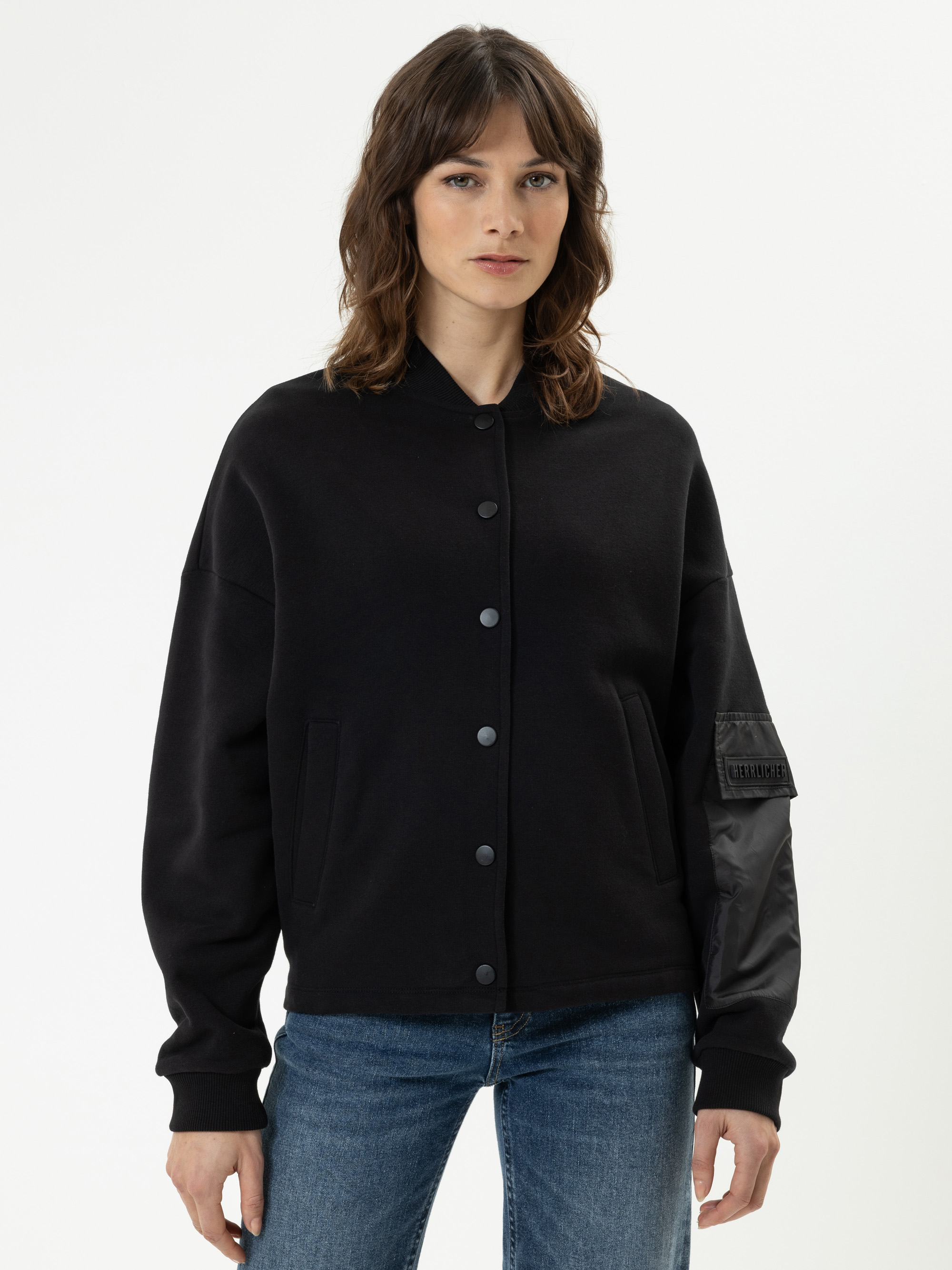 Eine Frau mit gewelltem braunem Haar trägt eine schwarze Jacke mit langen Ärmeln und blaue Jeans vor einem schlichten weißen Hintergrund.