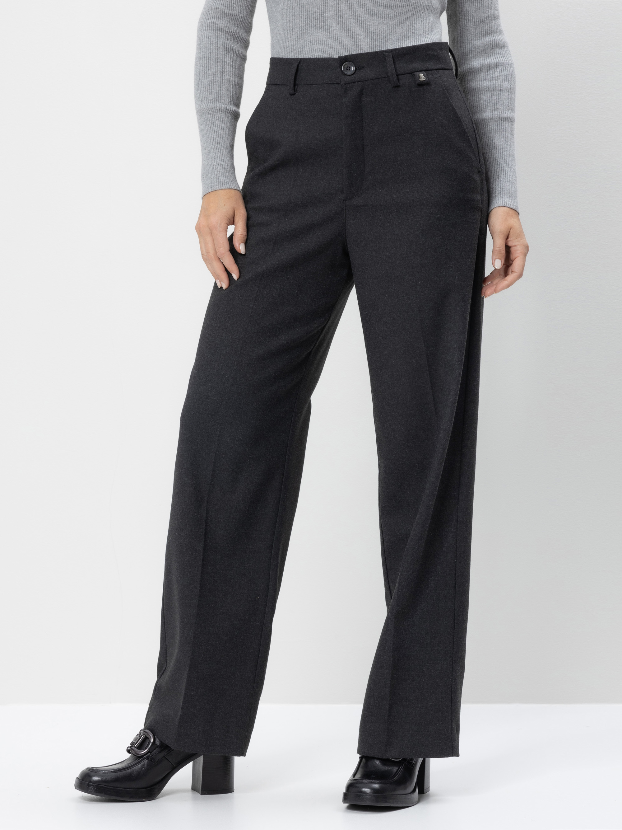 Eine Person, die eine dunkelgraue Hose mit weitem Bein und hoher Taille, ein hellgraues geripptes langärmeliges Oberteil und schwarze hochhackige Schuhe trägt, steht vor einem schlichten hellen Hintergrund. Ihre Hände befinden sich an der Seite.