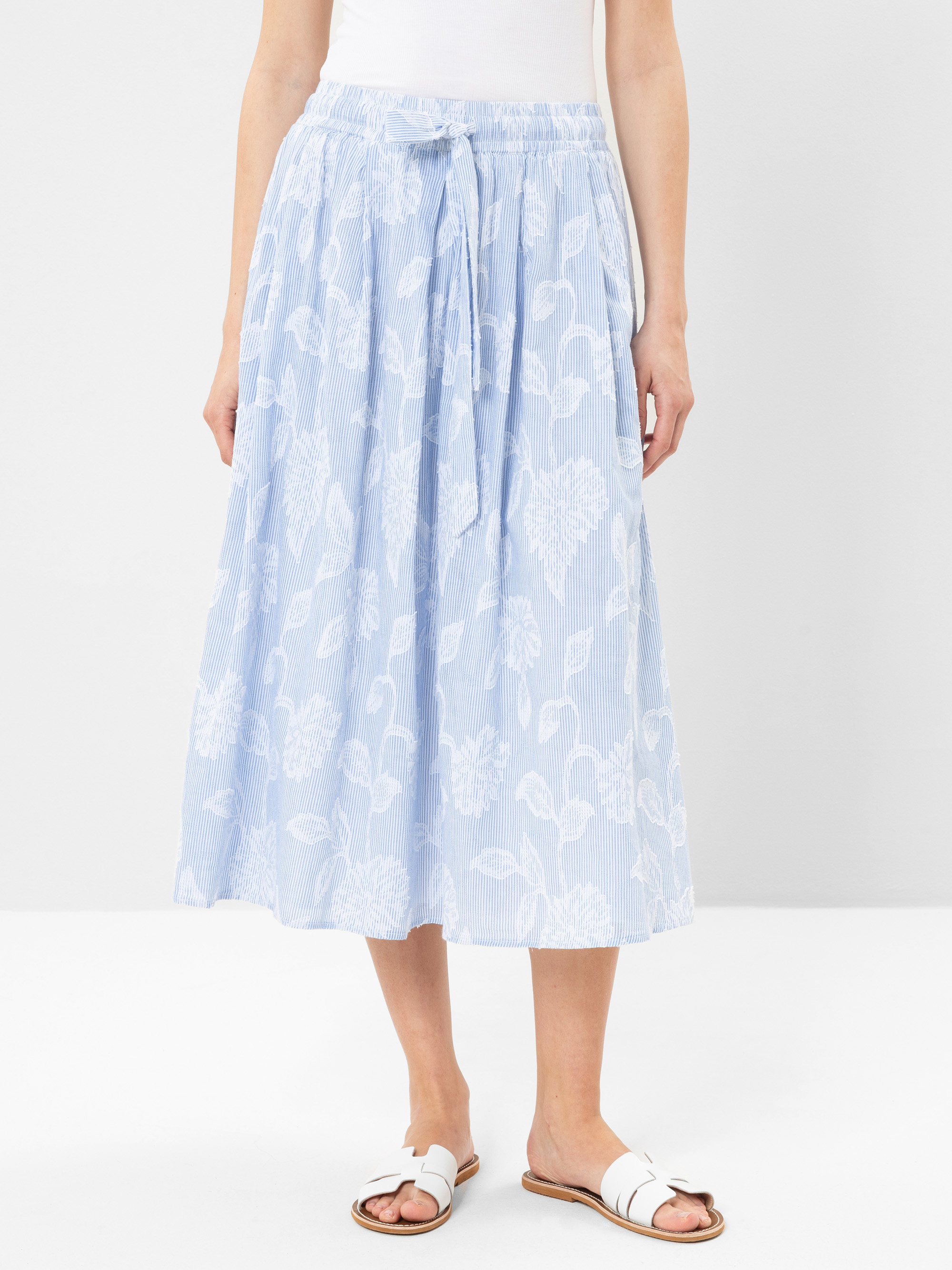 Eine Person, die einen hellblauen, blumengemusterten Midirock mit gebundener Taille, ein weißes Oberteil und weiße Sandalen trägt, steht vor einem schlichten weißen Hintergrund.