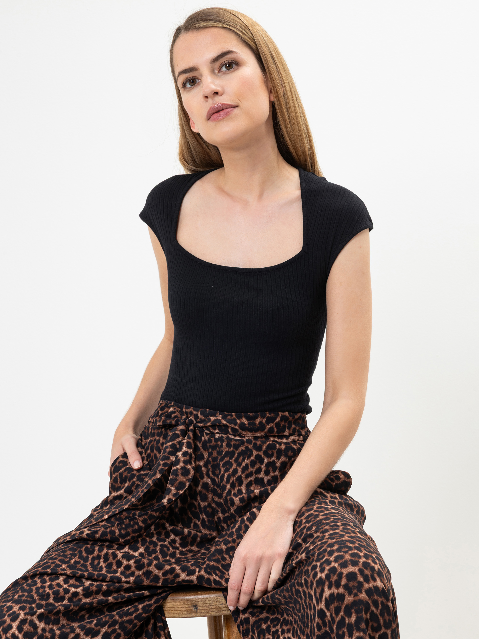 Eine Frau mit langem, glattem Haar trägt ein schwarzes, kurzärmeliges Oberteil und eine hoch sitzende Hose mit weitem Bein und Leopardenmuster. Sie sitzt auf einem Hocker vor einem schlichten weißen Hintergrund.
