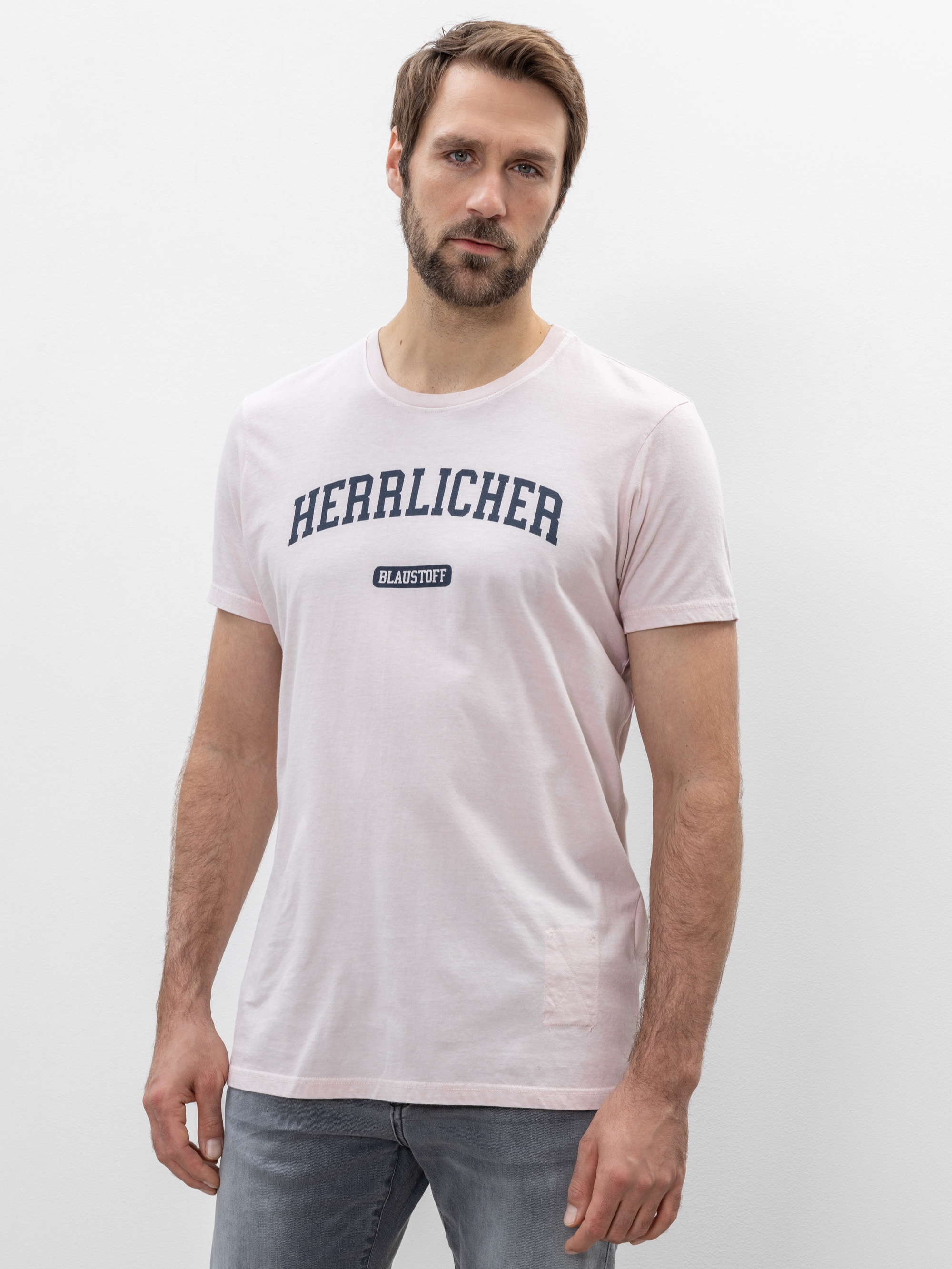 Ein Mann mit kurzen braunen Haaren und einem Bart trägt ein hellrosa T-Shirt mit der Aufschrift HERRLICHER und einem kleinen schwarzen Etikett mit der Aufschrift BLAILSTOFF auf der Vorderseite. Er steht vor einem schlichten weißen Hintergrund.