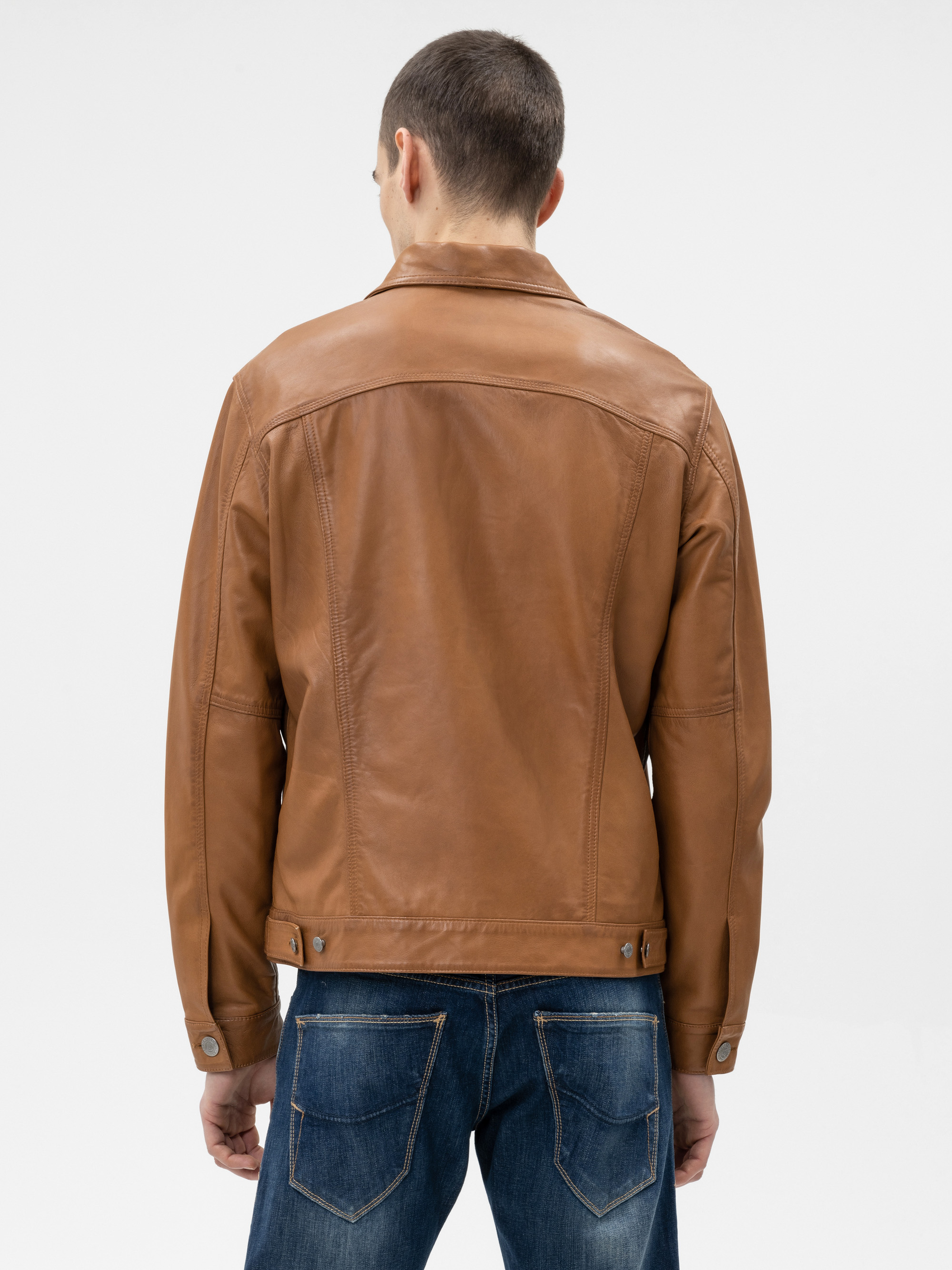 Von hinten ist ein Mann in hellbrauner Lederjacke und blauer Jeans zu sehen, der vor einem schlichten weißen Hintergrund steht.