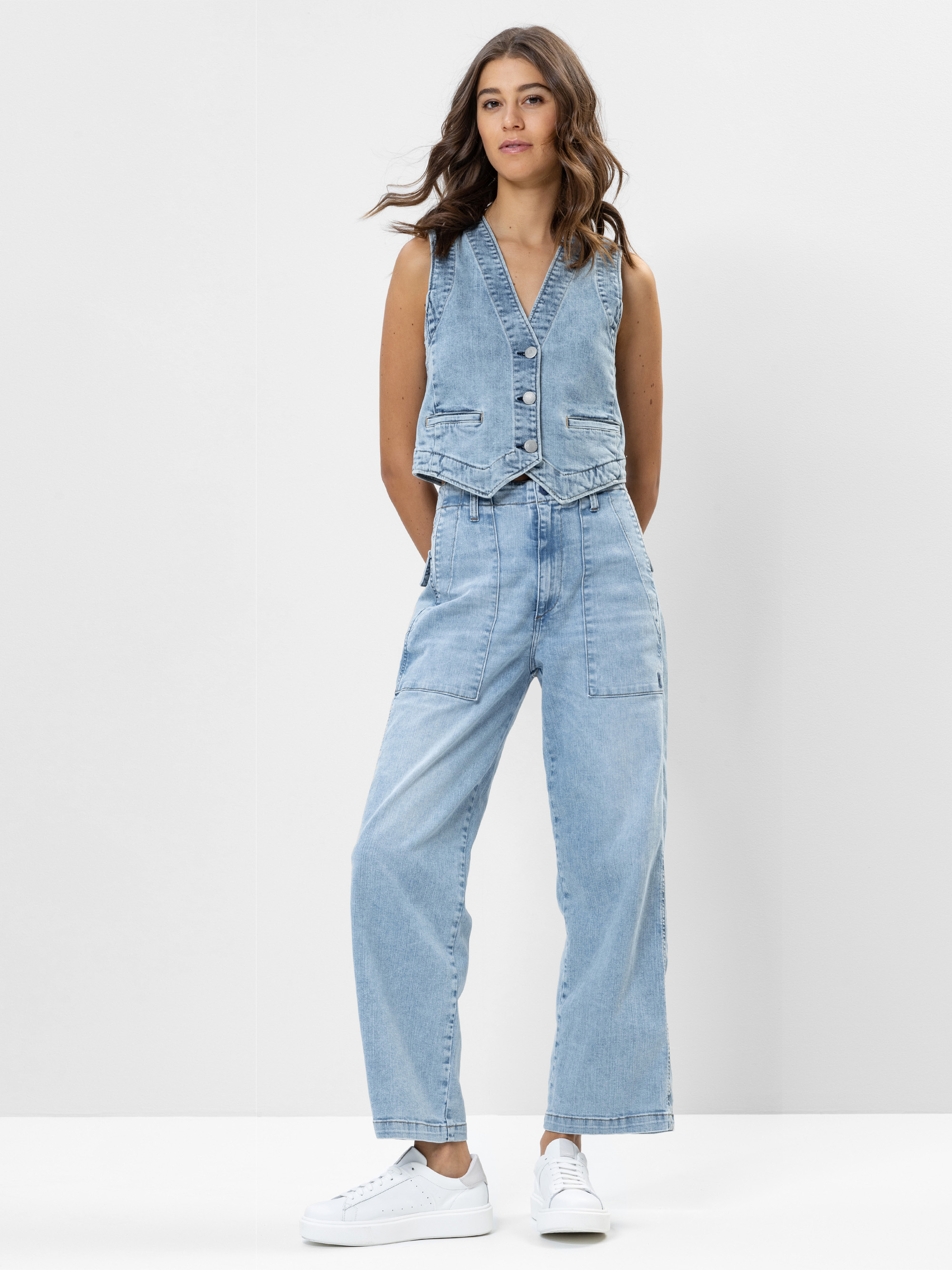 Eine Frau steht vor einem weißen Hintergrund und trägt eine ärmellose Jeansweste mit V-Ausschnitt und hellblaue Jeans mit hoher Taille und großen Taschen. Außerdem trägt sie weiße Turnschuhe und hat lockere, gewellte Haare.