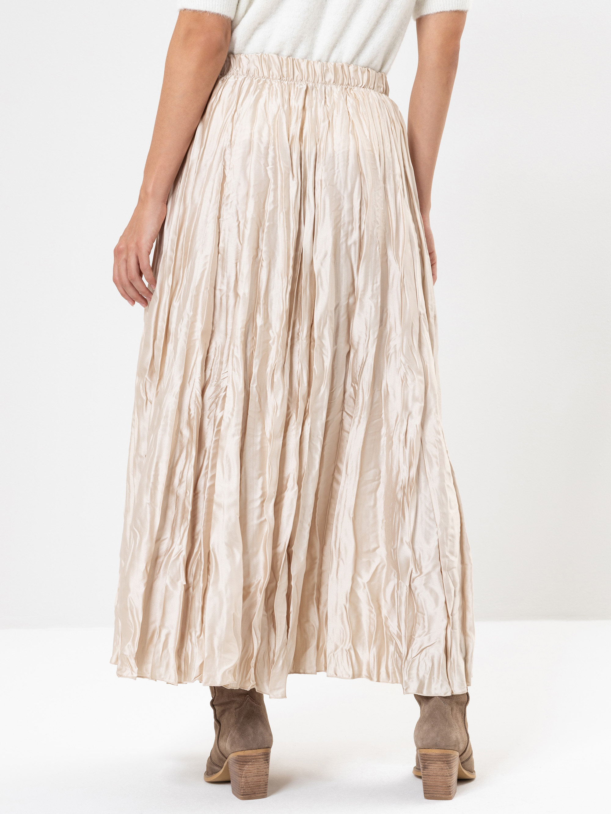 Eine Person trägt einen cremefarbenen, zerknitterten Maxirock mit elastischem Bund, gepaart mit taupefarbenen Ankle Boots. Die Person steht vor einem schlichten weißen Hintergrund, von hinten gesehen.