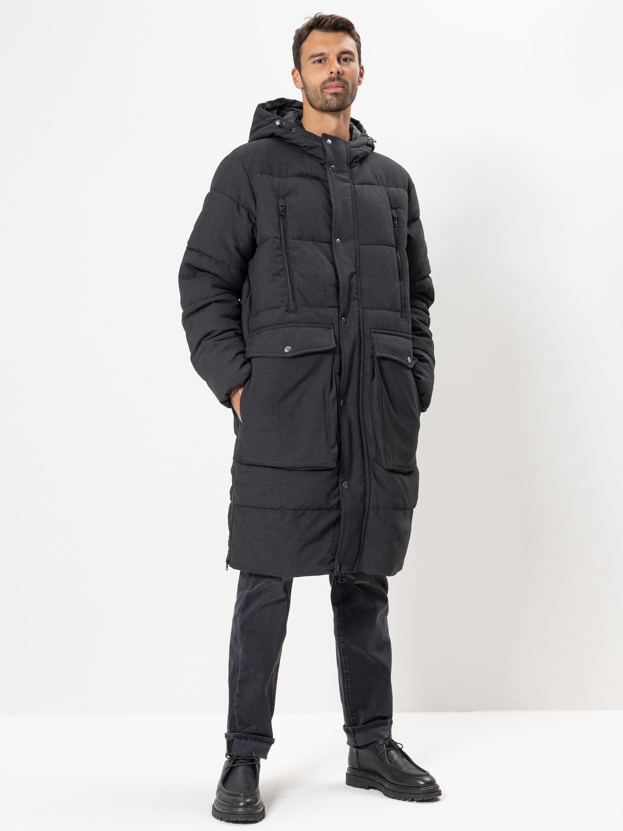 Ein Mann steht vor einem weißen Hintergrund und trägt einen langen, schwarzen Puffermantel mit großen Taschen, eine schwarze Hose und schwarze Schuhe. Er hat kurze Haare und einen Bart, einen selbstbewussten Gesichtsausdruck und die Hände in den Mante