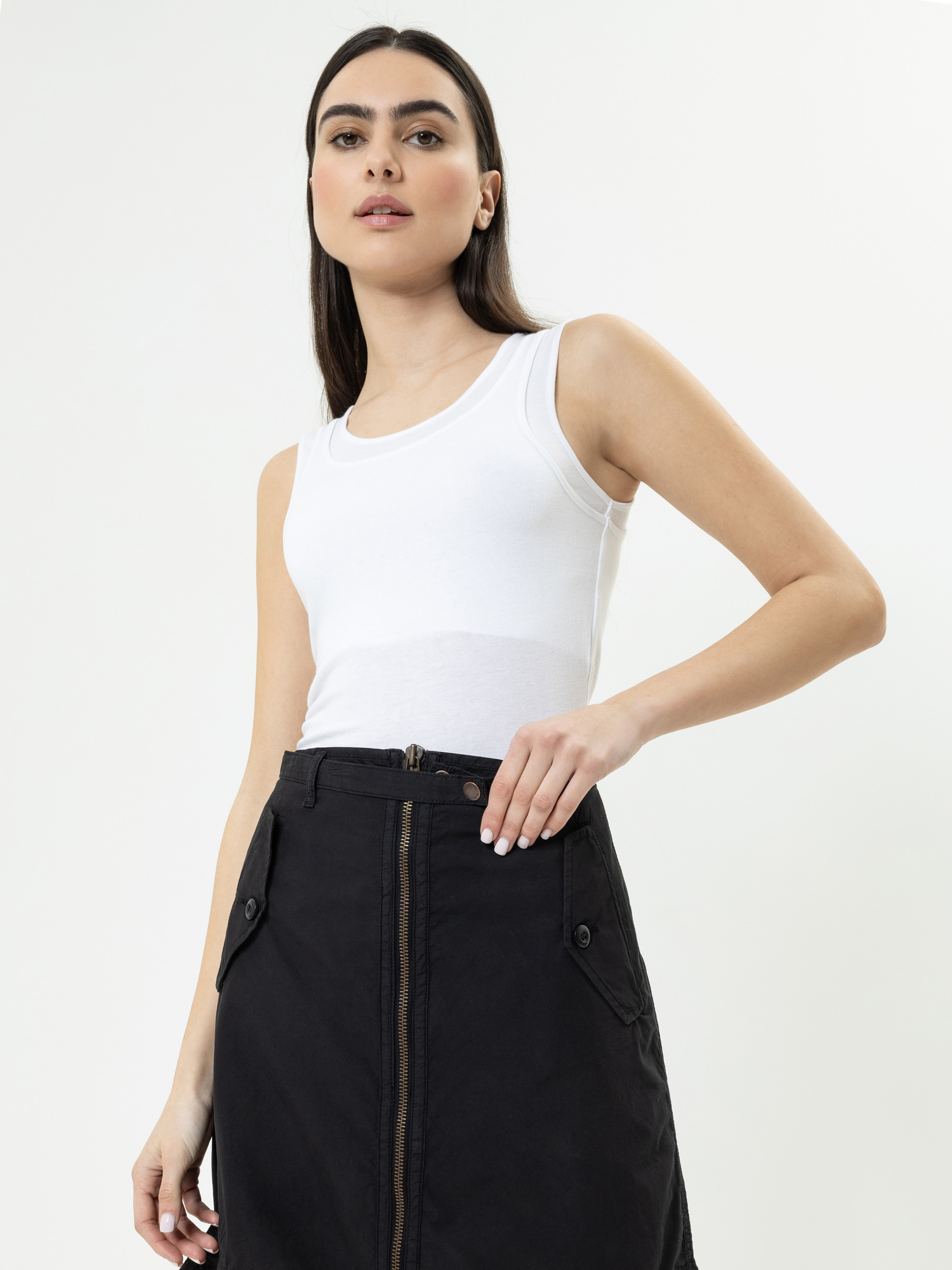 Eine Frau mit glattem dunklem Haar trägt ein weißes, ärmelloses Oberteil und einen schwarzen Rock mit hoher Taille und einem Reißverschluss vorne.