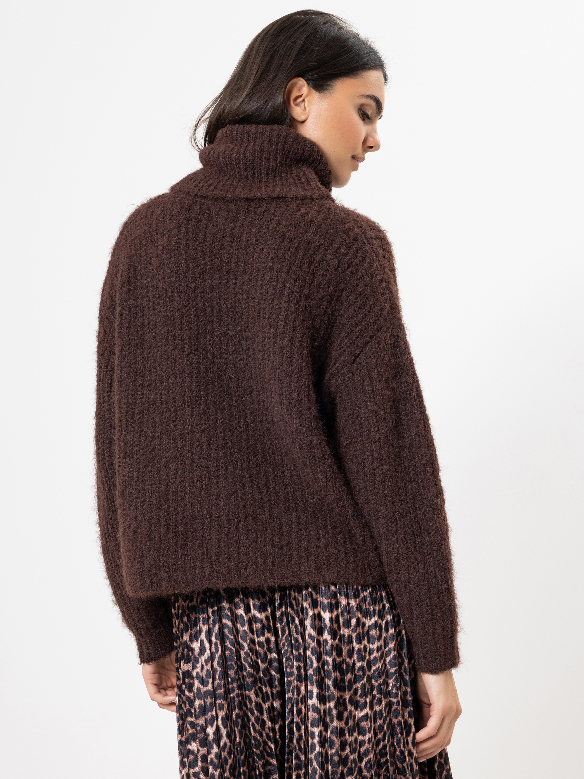 Eine Frau mit dunklen Haaren, die einen dicken braunen Rollkragenpullover und einen Rock mit Leopardenmuster trägt, steht mit dem Gesicht zur Seite und blickt über ihre Schulter auf einen schlichten weißen Hintergrund.