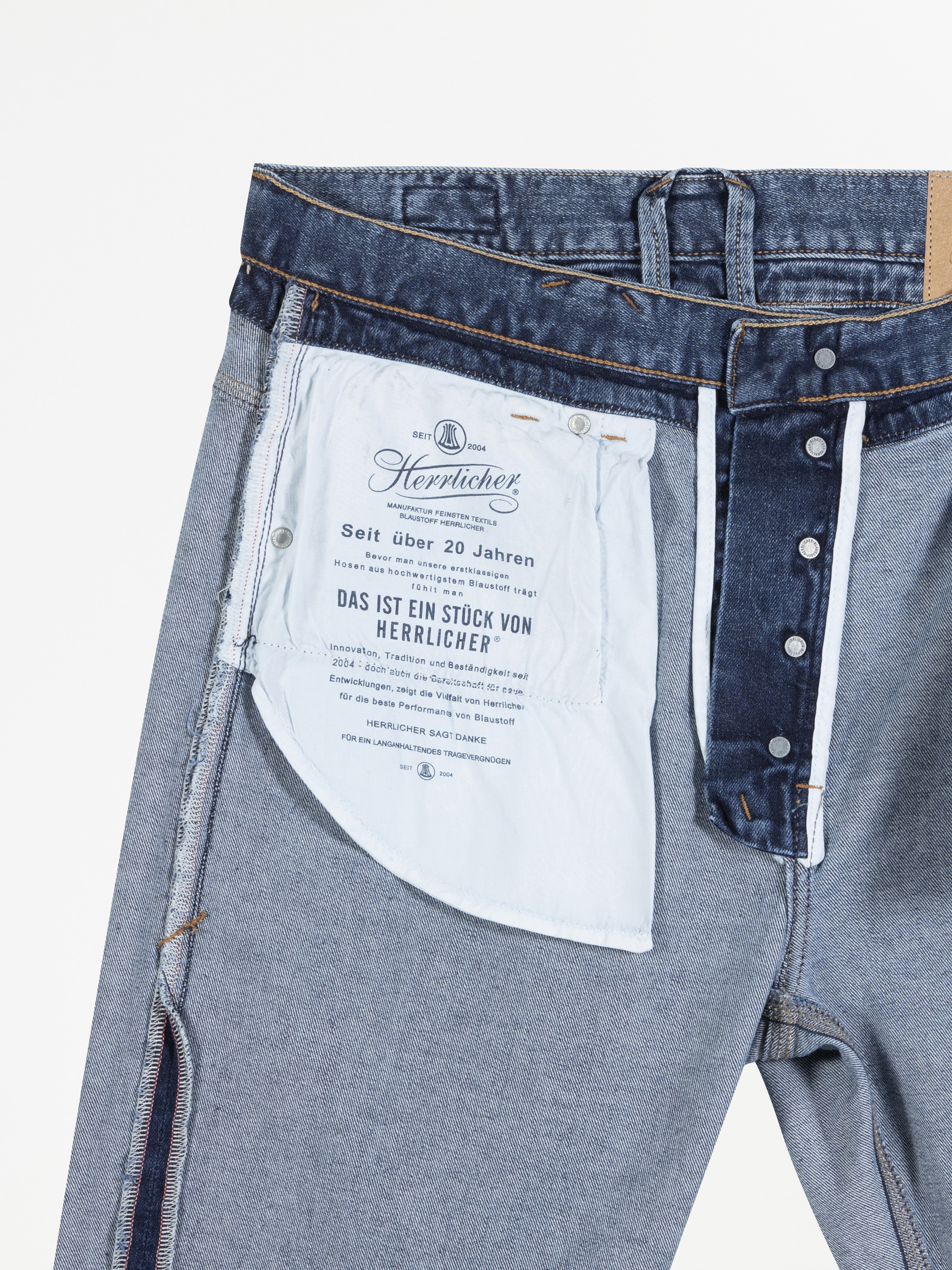 Eine blaue Jeans wird auf links gedreht und teilweise aufgeknöpft, so dass ein weißes Taschenfutter mit aufgedruckten deutschen Texten und Logos zum Vorschein kommt. Die Nähte und Denim-Details sind deutlich sichtbar.