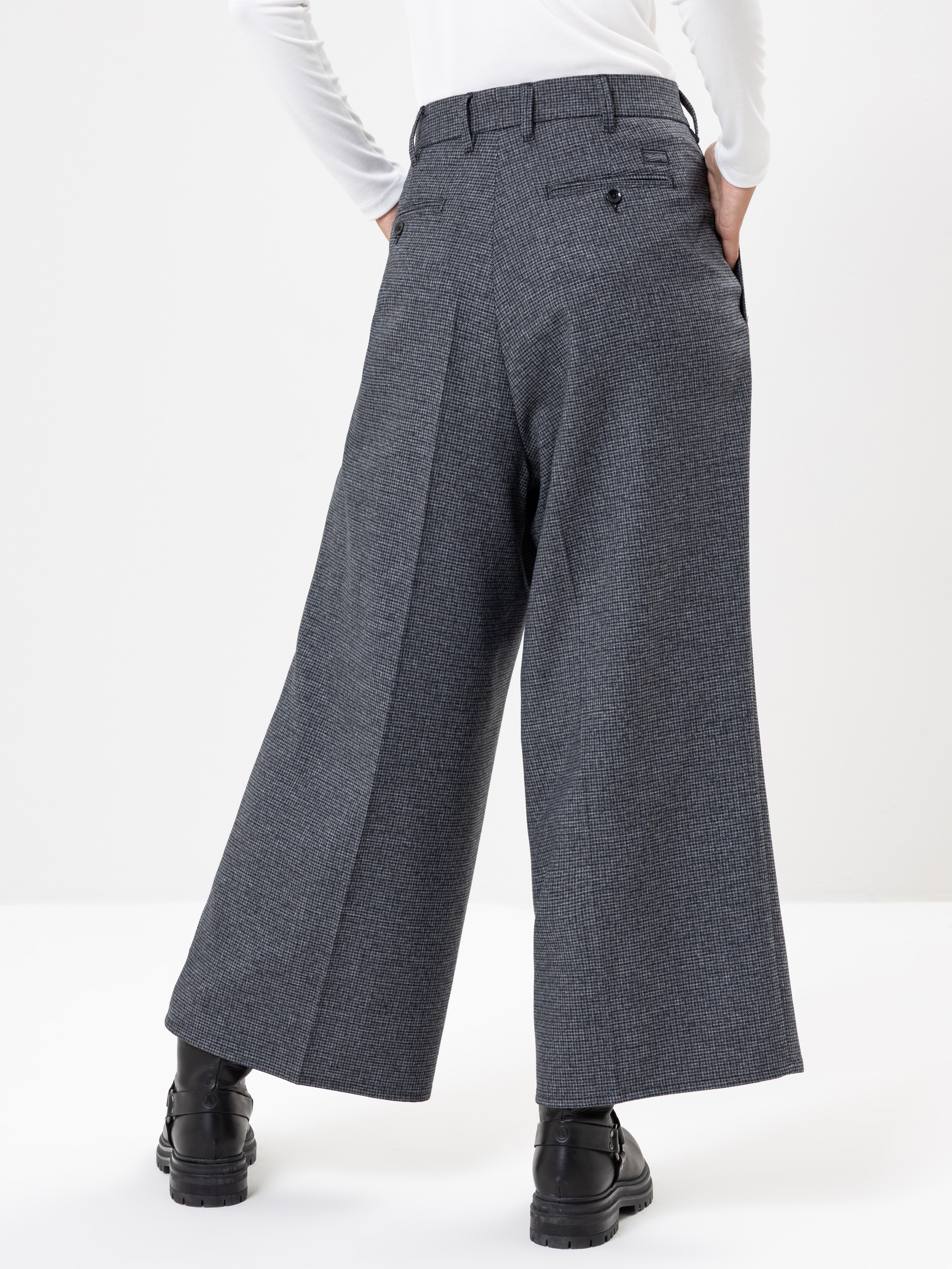 Person, die eine graue Hose mit weitem Bein und hoher Taille, Gesäßtaschen und schwarzen Schuhen trägt. Sie steht mit den Händen in den Hosentaschen und schaut von der Kamera weg vor einem schlichten weißen Hintergrund.