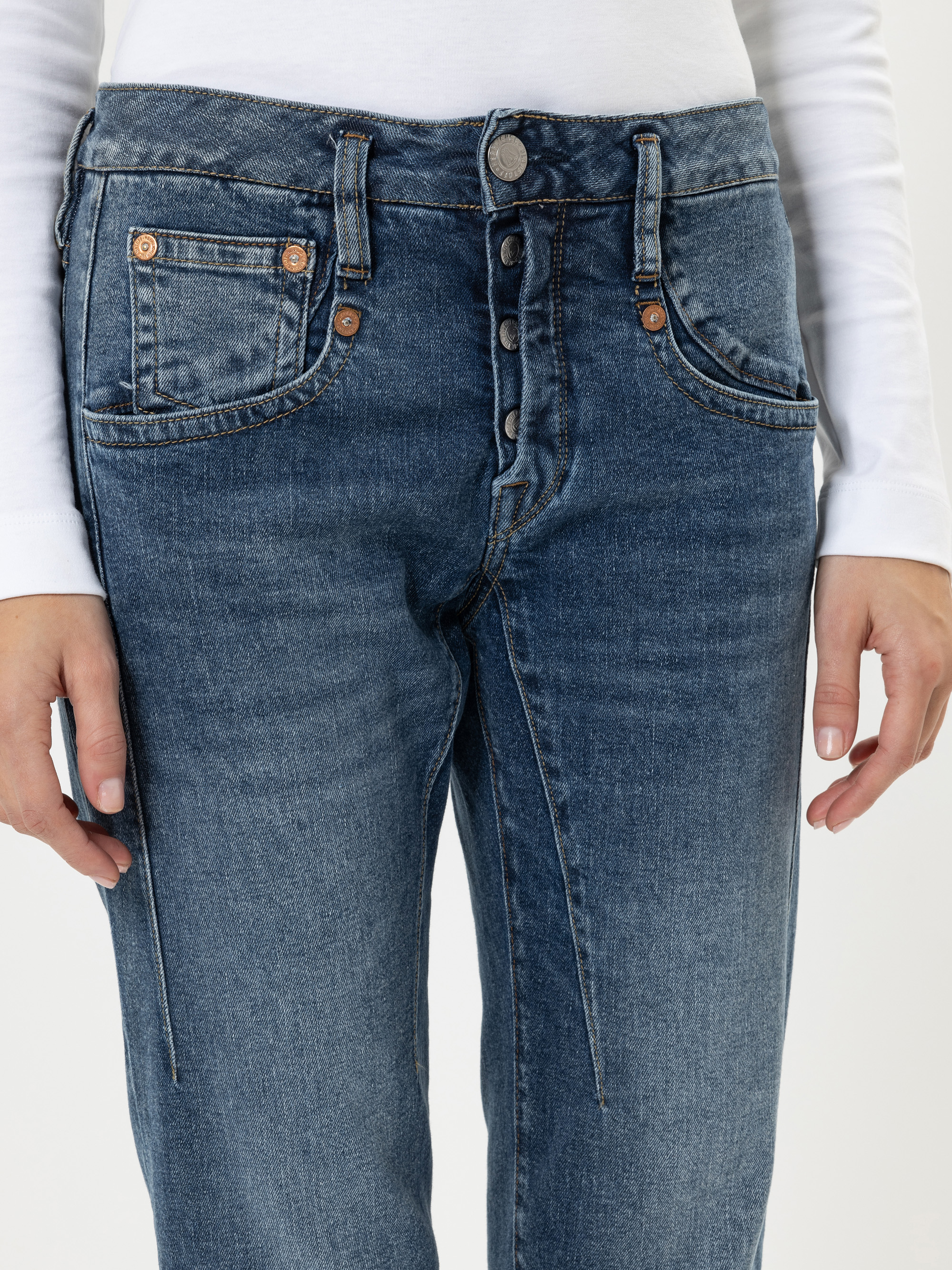 Eine Person, die ein weißes, langärmeliges Hemd und eine blaue, hochgekrempelte Jeans mit sichtbarem Knopfverschluss und Vordertaschen trägt. Das Bild ist so beschnitten, dass nur der Torso und die Oberschenkel zu sehen sind.