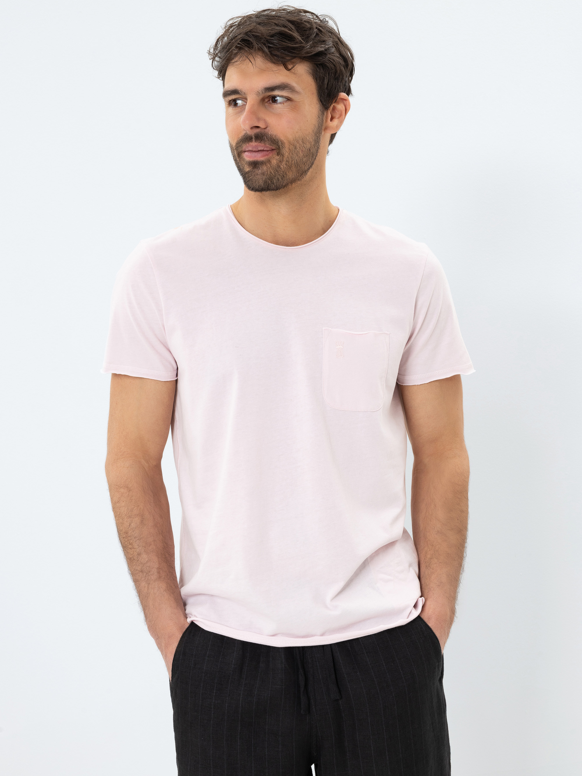 Ein Mann mit dunklem Haar und Bart, der ein hellrosa T-Shirt mit Brusttasche und eine schwarze Hose trägt, steht vor einem schlichten weißen Hintergrund und schaut leicht zur Seite, die Hände in den Taschen.