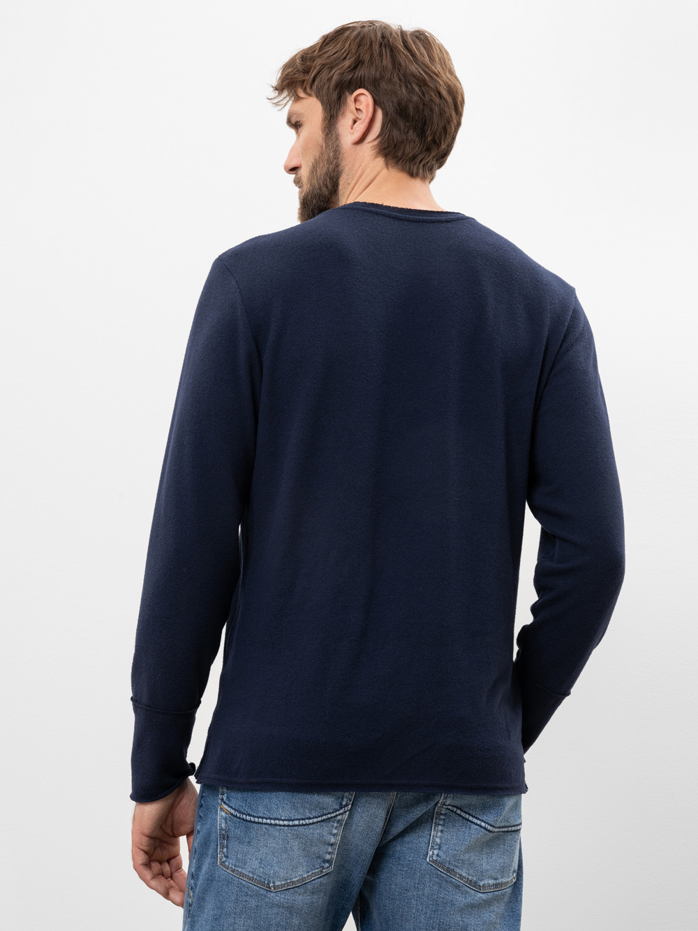 Man sieht einen Mann mit kurzen braunen Haaren von hinten, der ein marineblaues langärmeliges Hemd und blaue Jeans trägt und vor einem schlichten weißen Hintergrund steht.