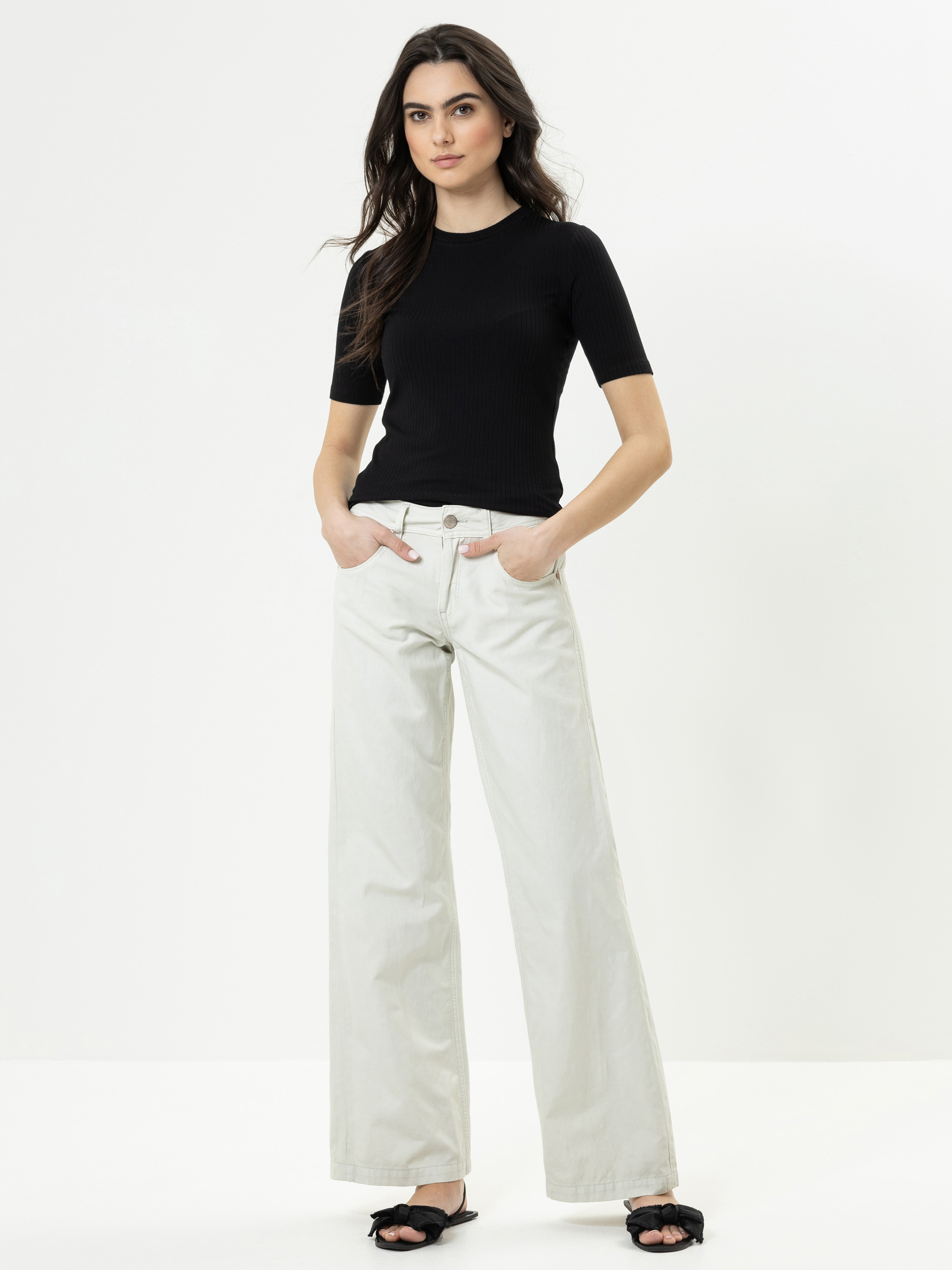 Eine Frau steht vor einem einfarbig weißen Hintergrund und trägt ein schwarzes, kurzärmeliges Oberteil, helle, weit geschnittene Jeans und schwarze, hochhackige Sandalen mit offenen Zehen. Sie hat langes dunkles Haar und die Hände in den Taschen.