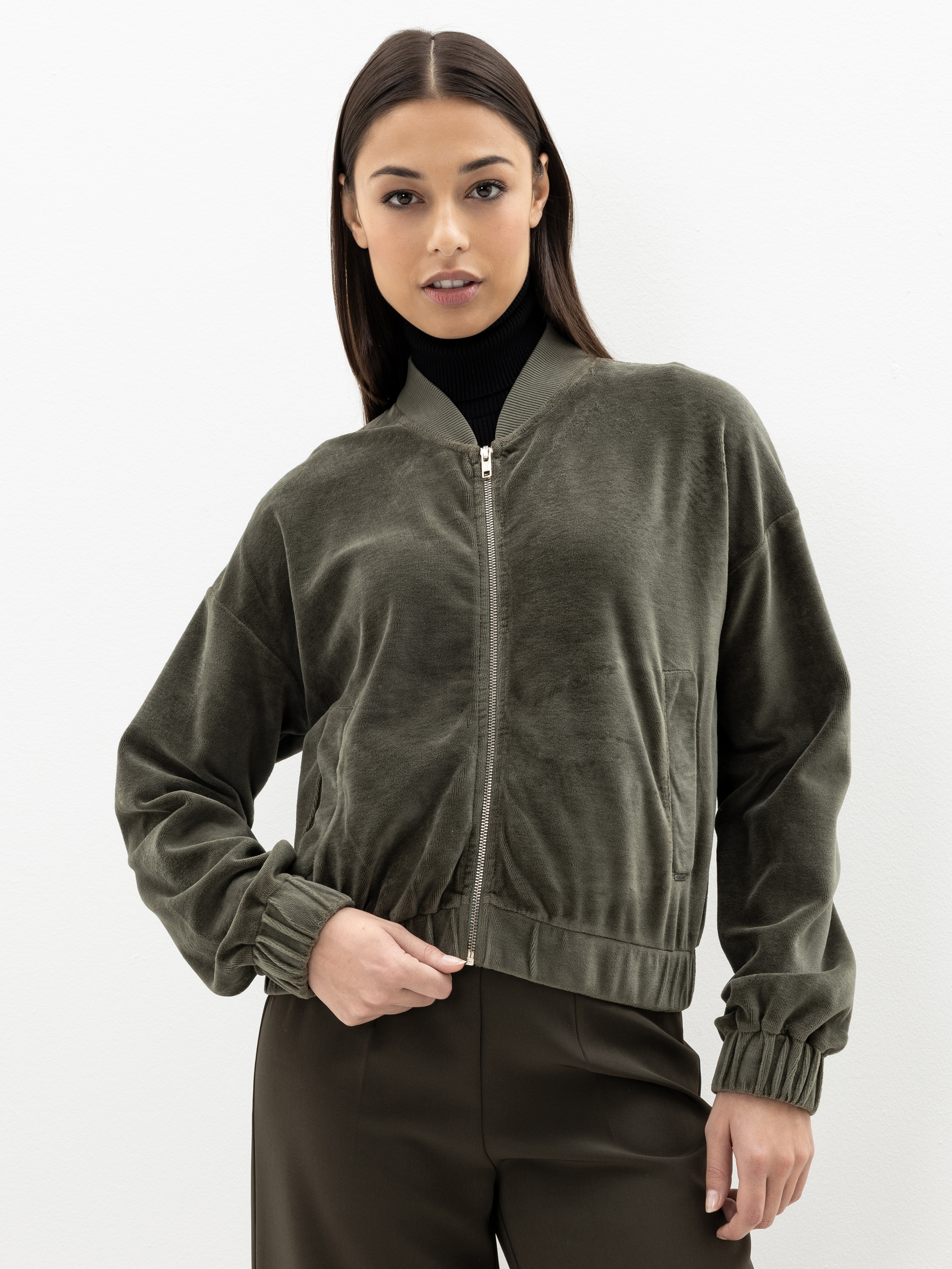 Eine Frau, die eine olivgrüne Bomberjacke mit Reißverschluss über einem schwarzen Rollkragenpullover trägt, steht vor einem schlichten weißen Hintergrund, eine Hand in der Tasche und mit entspannter Miene.