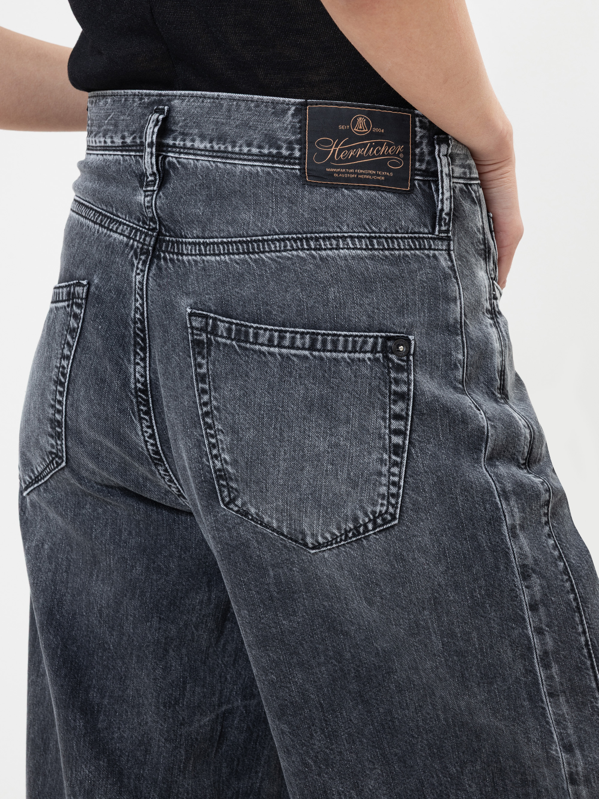 Eine Person, die eine hoch taillierte, locker sitzende, verwaschene schwarze Jeans mit einem sichtbaren Herrlicher-Markenaufnäher am Bund trägt, steht vor einem einfarbig weißen Hintergrund.