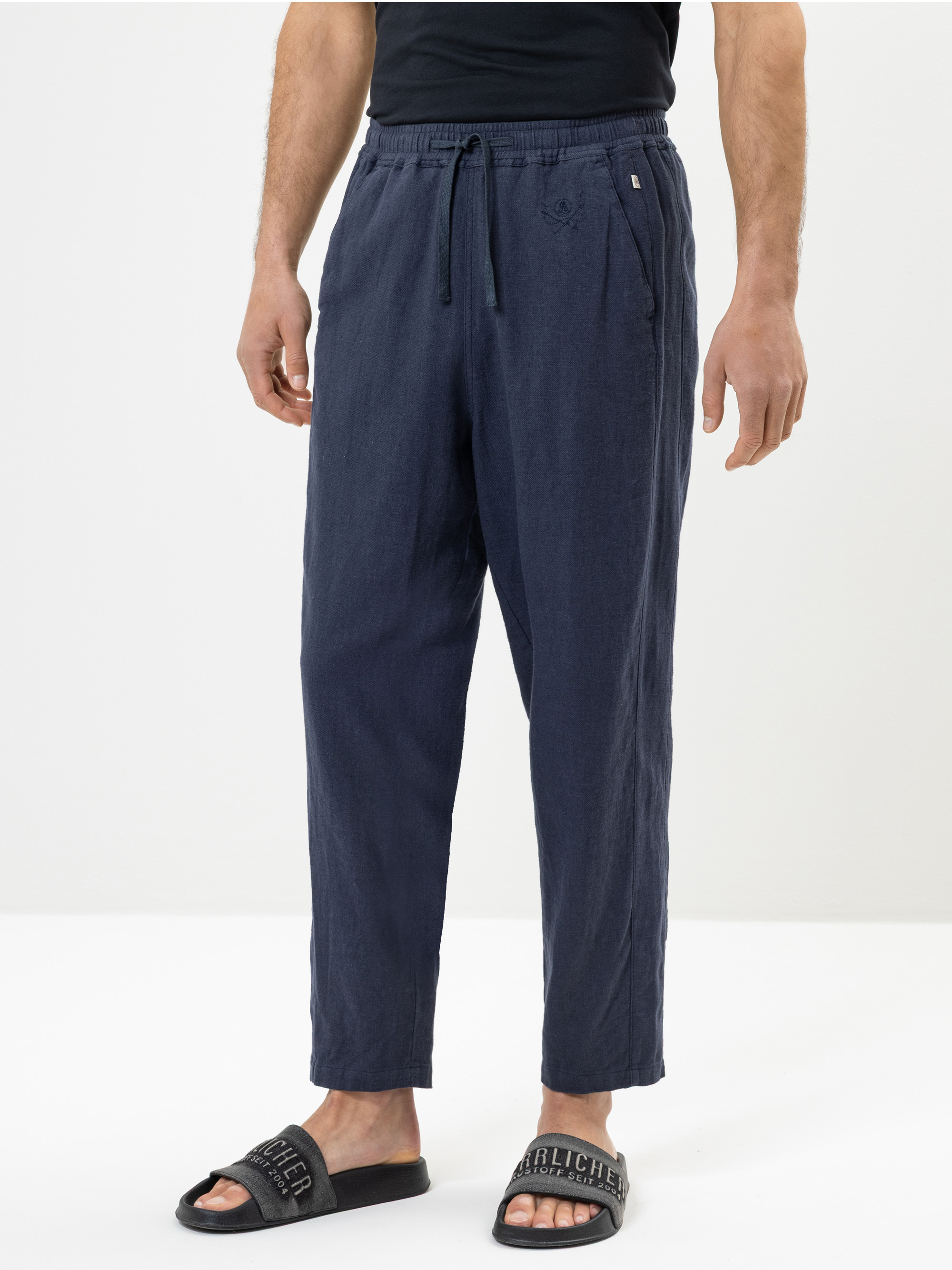 Eine Person, die eine marineblaue Hose mit Kordelzug, ein schwarzes T-Shirt und schwarze Slide-Sandalen mit weißer Aufschrift trägt, steht vor einem schlichten weißen Hintergrund. Das Foto zeigt die Figur von den Schultern bis zu den Füßen.