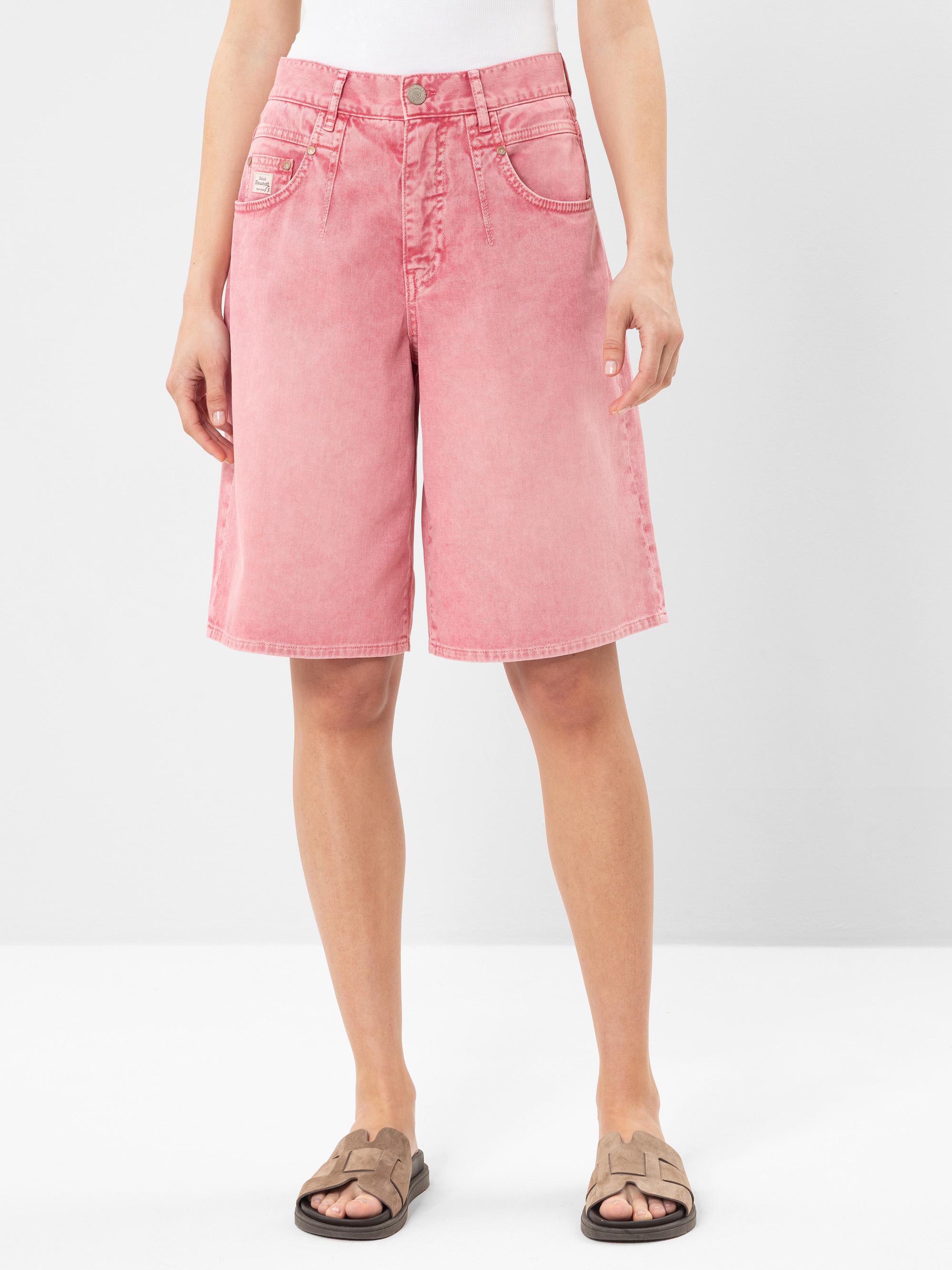 Eine Person, die hochtaillierte, rosafarbene Bermuda-Shorts aus Jeansstoff, ein weißes Oberteil und taupefarbene Slide-Sandalen trägt, steht vor einem schlichten weißen Hintergrund. Die Shorts haben Seiten- und Gesäßtaschen mit sichtbaren Nähten.