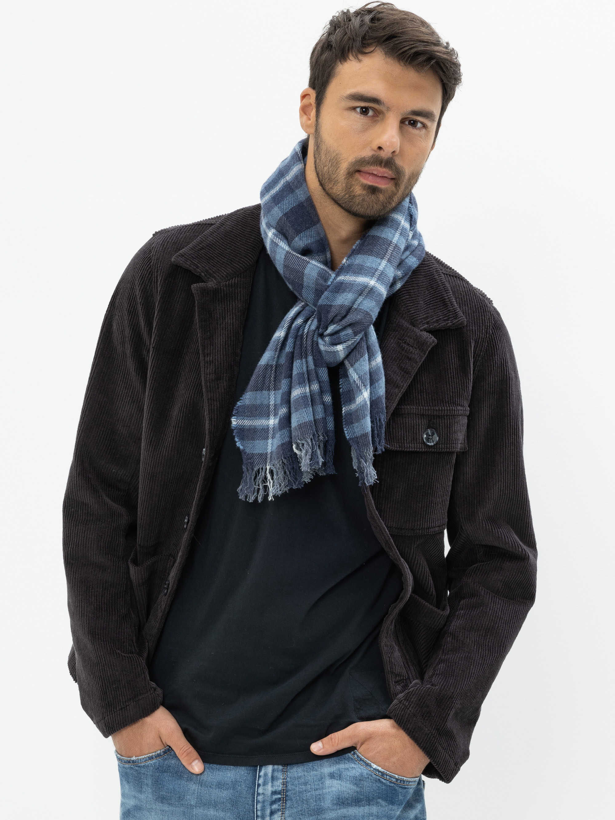 Ein Mann mit kurzen dunklen Haaren und Bart trägt eine dunkle Kordjacke, ein schwarzes Hemd, einen blau karierten Schal und blaue Jeans. Er steht mit den Händen in den Taschen vor einem schlichten weißen Hintergrund.